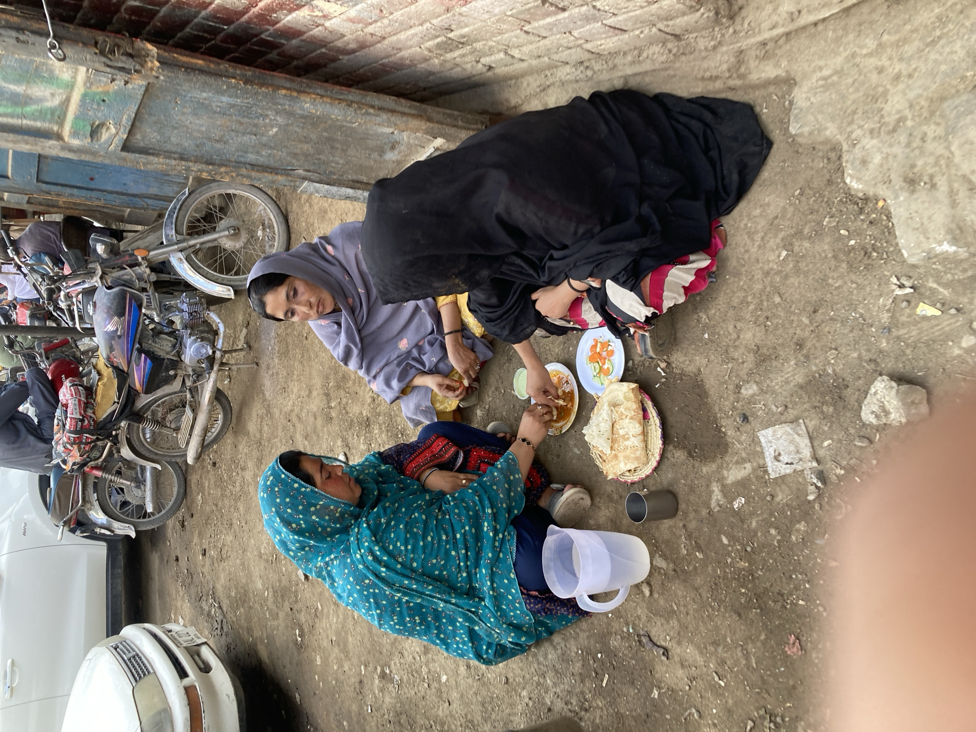 This image captures three women sharing a meal outdoors on a rough, dirt ground in what appears to be an alleyway or a roadside area. The scene is set during daylight hours, under natural light.
Two women are seated on the ground near the center of the frame, surrounded by food. One woman wears a light grey head covering and matching attire, looking towards the food. Beside her, another woman, mostly covered by a black head covering (possibly a niqab or burqa), is actively taking food from a small white plate containing a curry or stew. Adjacent to this, there's another plate with sliced vegetables or salad. A basket filled with flatbread (likely naan or roti) is placed near the food items.
A third woman, dressed in a vibrant teal headscarf with a floral pattern and matching clothes, is reclining on the ground further to the right. A clear plastic pitcher, seemingly filled with a white liquid, and a metal cup are placed near her.
In the background, on the left side of the frame, several motorcycles are parked closely together, with their handlebars and front wheels prominently visible. The front bumper and headlights of a white car are also partially visible on the far left. Above the parked vehicles, parts of a brick wall and a weathered wooden beam or structure can be seen, indicating the proximity of a building.
The women appear to be engaged in a communal meal or a break. Their expressions are calm and focused on the activity. The visible text on the white car's license plate is partially obscured but contains numbers such as "02 2777" and letters that might spell "ALL". Other text on the motorcycles is not clearly legible. The overall setting suggests a casual, outdoor gathering in a less formal environment.