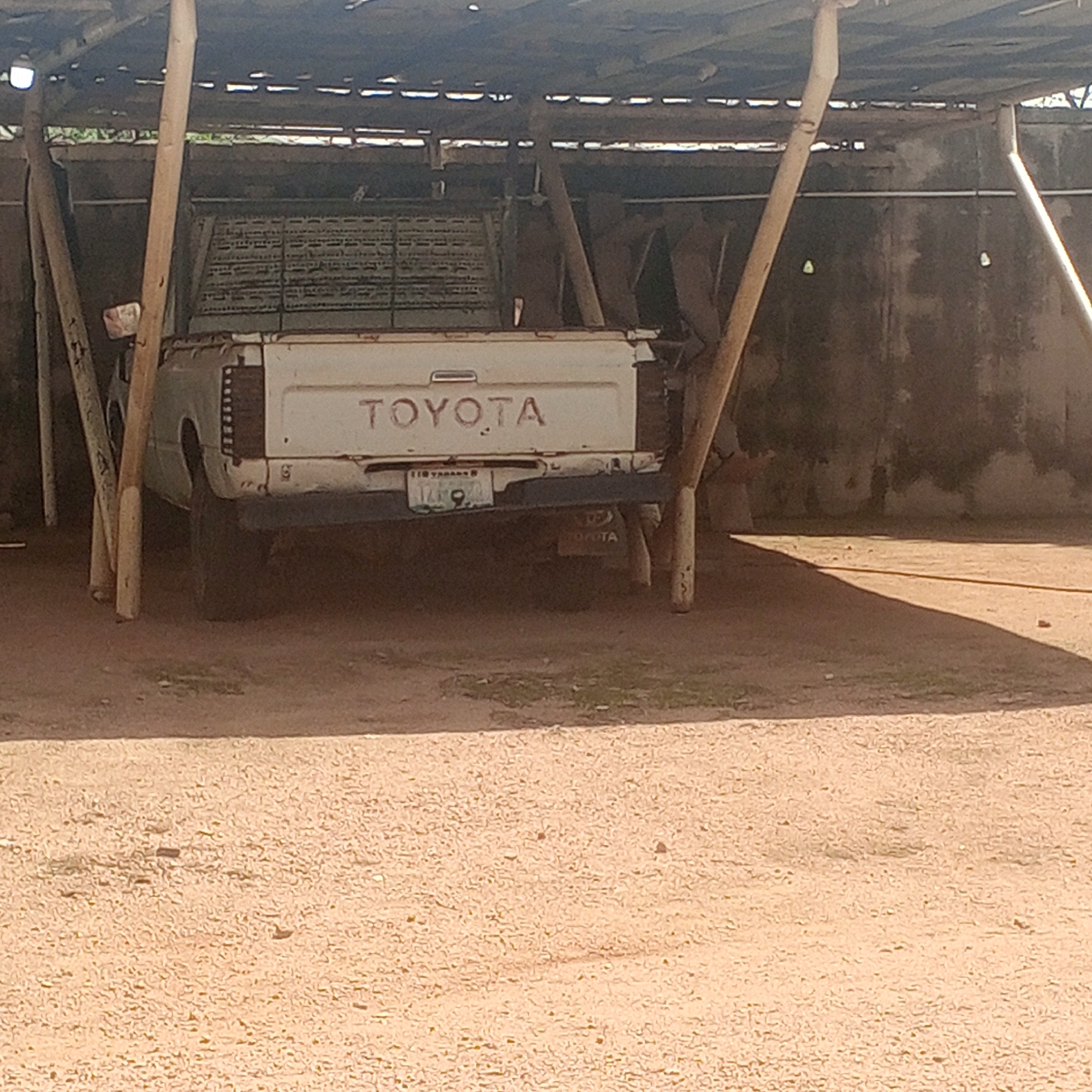 A white Toyota pickup truck is parked under a rustic, open-air shelter supported by wooden posts. The truck's tailgate displays the "TOYOTA" logo and a license plate with an illegible number. The shelter's roof appears to be corrugated metal or similar material, with visible support beams and some openings. The ground beneath and around the truck is covered in reddish-brown dirt and small stones. In the background, a weathered concrete wall is visible. The scene is located in Zinna, Nigeria, suggesting a typical setting for vehicles in a rural or semi-urban environment.