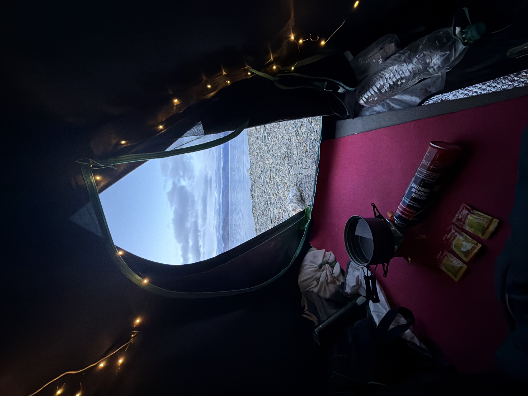 Inside a dark tent, a maroon sleeping mat is laid out. On the mat, a camping stove with a pot of water is set up. Next to the stove are three small packets of what appear to be seasonings or sauces, and a red propane canister. To the left of the stove, a bundle of fabric is visible. Further to the right, a plastic water bottle and other camping gear are present. Decorative string lights with yellow bulbs are draped along the tent's interior. The tent opening reveals a view of a body of water, a pebbly shoreline, and distant mountains under a cloudy sky. The location is identified as Karzok, India, suggesting a campsite near the shore of a lake or river in that region. No people are depicted in the image.