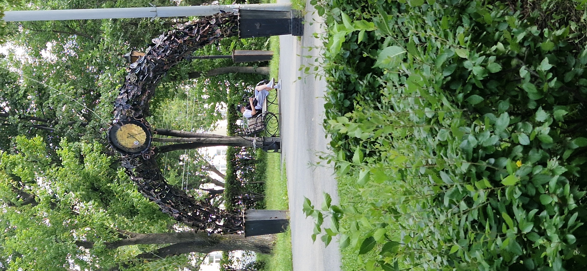 The image depicts a park scene in Miass, Russia. An ornate archway, adorned with metalwork resembling leaves and butterflies, spans a paved pathway. Atop the arch is a large, circular clock with Roman numerals. To the right of the arch is a tall, slender lamppost. A person wearing a white t-shirt and grey shorts is seated on a bench beneath the arch, looking down at a device in their hands. They are partially obscured by the foliage in the foreground. The environment is lush with green trees and bushes, suggesting a warm season. In the background, buildings can be glimpsed through the trees, indicating an urban setting. The ground is paved in the foreground, transitioning to grass and then more dense vegetation.