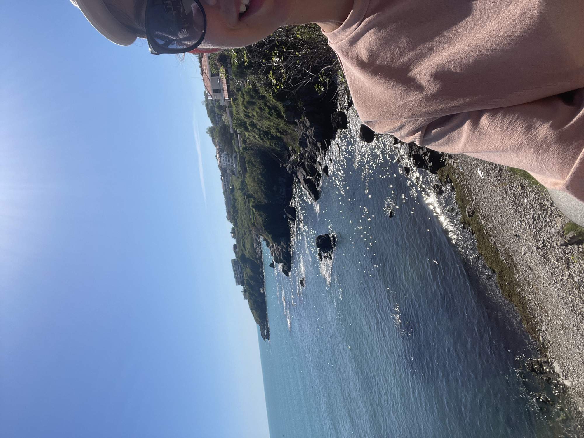 A sunny, rotated selfie shows a person wearing sunglasses and a beige top standing near a coastal cliff. The blue sea is visible below. The cliff is covered in green vegetation and some buildings, suggesting a residential area. A rocky shoreline is present along the water's edge. The location appears to be Sozopol, Bulgaria, given the coastal landscape and architectural style.