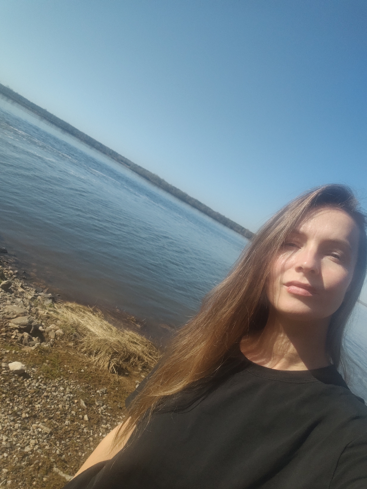 A woman with long brown hair and a black t-shirt stands near the shore of a body of water. The water is calm and stretches towards a distant treeline under a clear blue sky. The shoreline consists of small rocks, pebbles, and patches of dry grass. The picture may be taken in Chernushka, Russia.