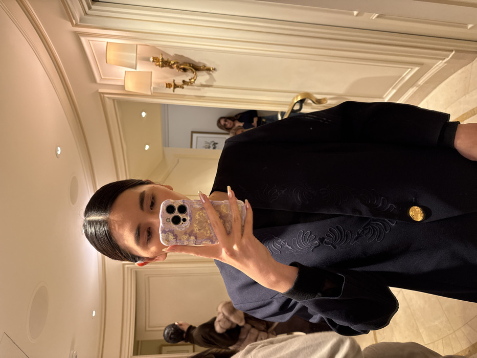 A person takes a selfie in a mirrored room. They hold a phone with a patterned case, displaying light-colored nails. The individual wears a dark blue blazer with decorative embroidery and a prominent gold button. In the reflection, another person stands in a doorway. The room features ornate wall sconces and detailed moldings. Other people are visible in the mirror.