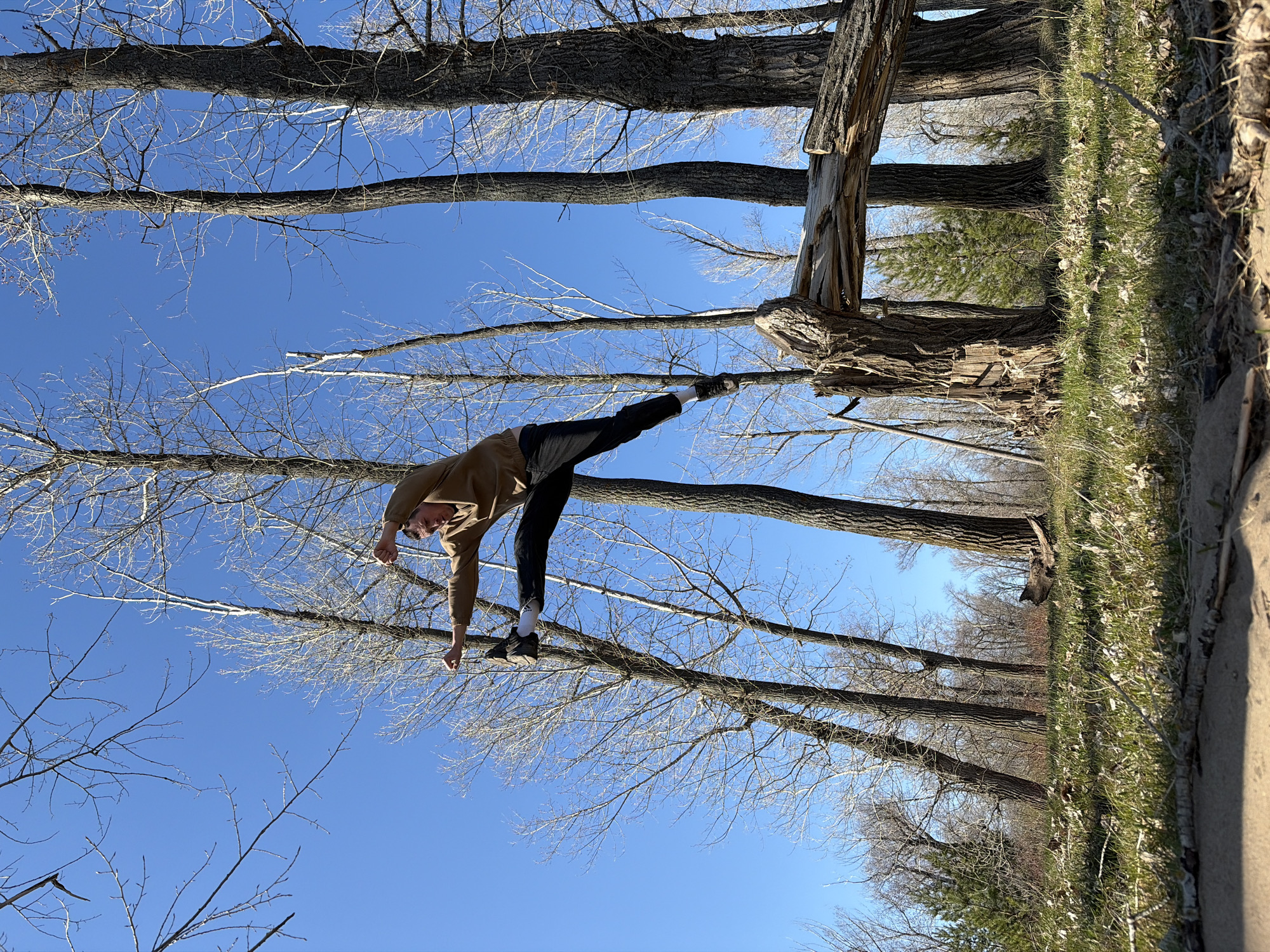 A man is performing a backbend in a wooded area, likely a park. The trees are mostly bare of leaves. The sky is blue. The man is wearing a tan jacket, black pants, and black and white sneakers. He is bending backward, with one arm reaching towards the ground and one leg raised. The ground is covered in grass. The location is not definitively identifiable as Chaykovsky, Russia, based on the visual information provided.