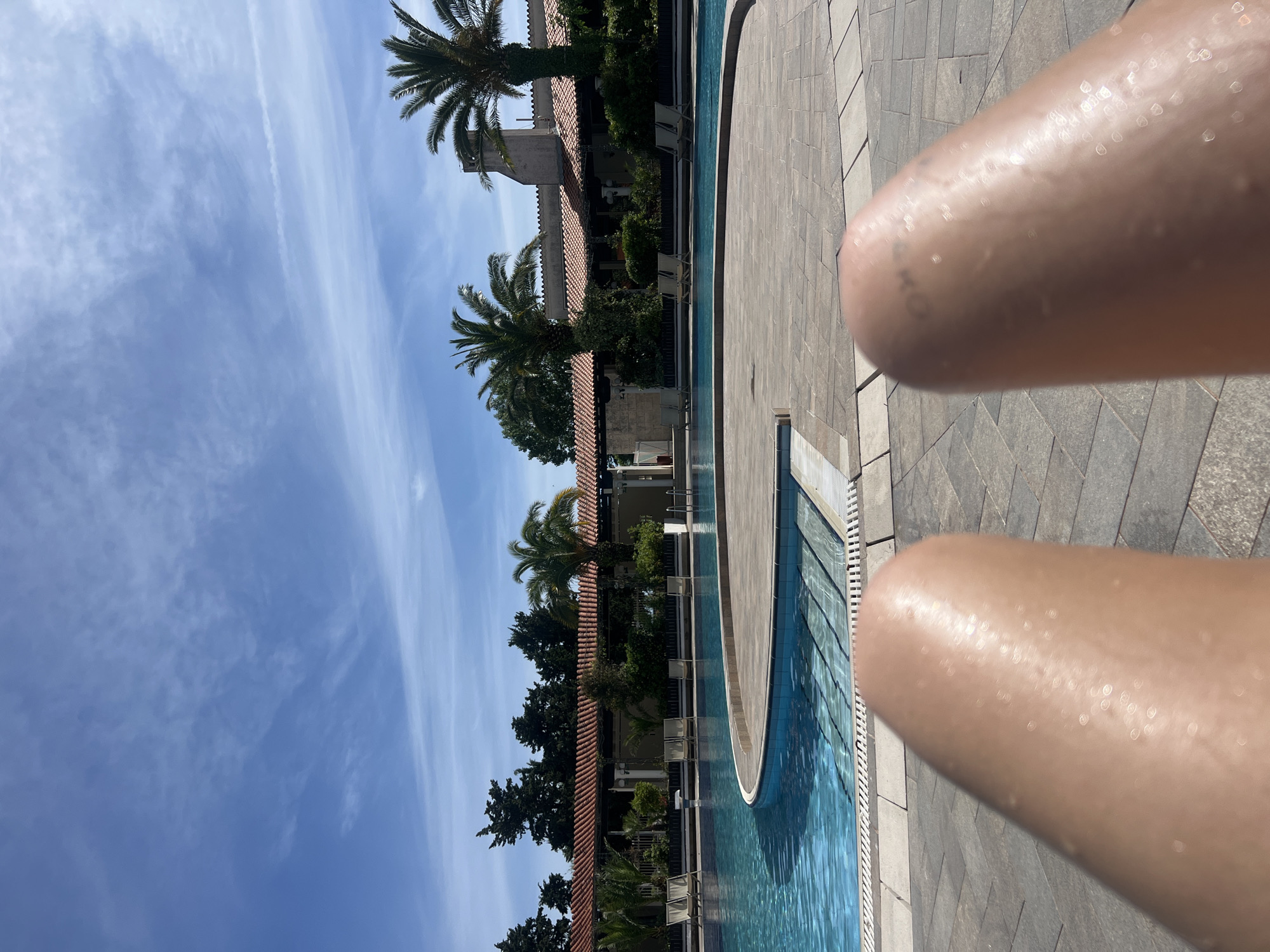 A sunny daytime image captures a poolside scene at a resort in Budva, Montenegro. The composition includes a portion of a swimming pool with clear blue water, adjacent to a tiled pool deck. In the foreground, a partial view of a person's legs is visible, covered in water droplets. The background features palm trees and a building with a terracotta-tiled roof. The sky is blue with wispy clouds.