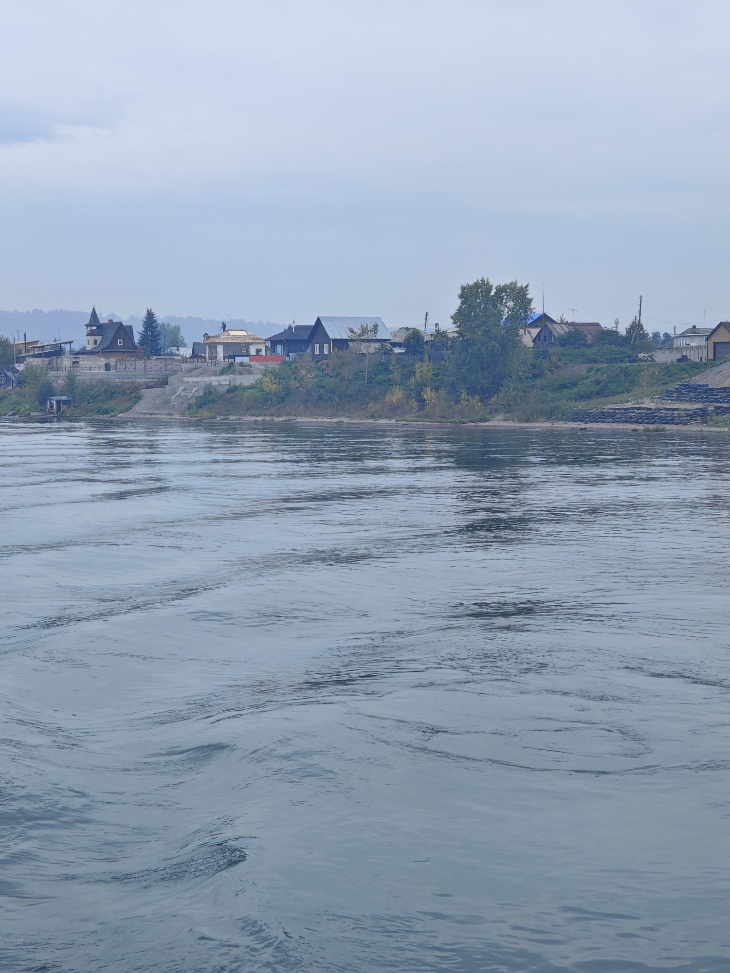 The image depicts a wide, calm river with gentle ripples, possibly the Yuryuzan River in Ermolaevo, Russia. On the far bank, a settlement of houses is visible, situated on a slightly elevated terrain. The houses are varied in style and size, with some appearing to be under construction, indicated by visible scaffolding and roofing work. Green vegetation, including trees and bushes, surrounds the houses and lines the riverbank. The sky is overcast and a muted gray, contributing to the overall subdued lighting of the scene. The foreground is dominated by the water, with the settlement providing a backdrop.