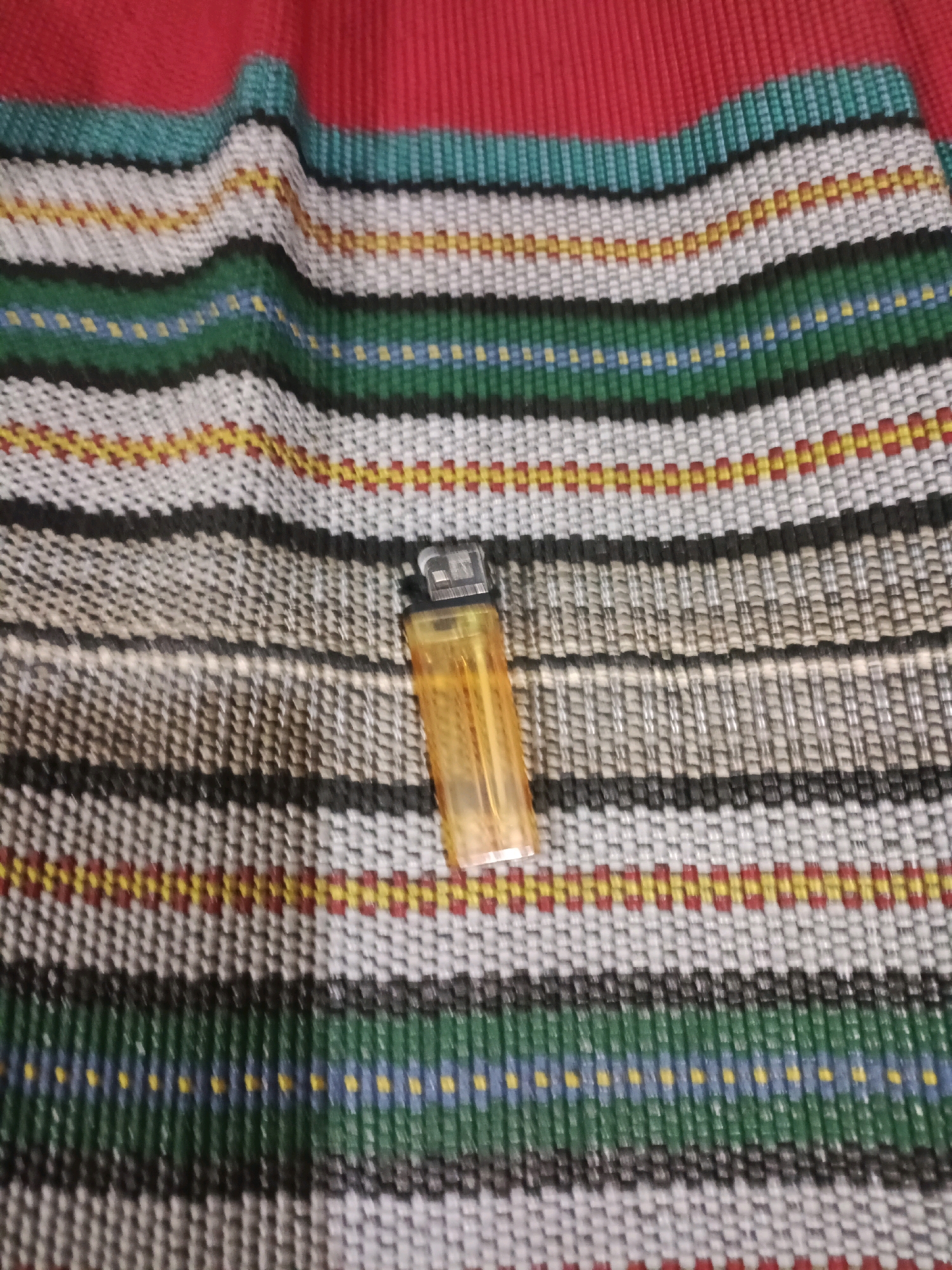 An orange disposable lighter rests on a woven placemat with horizontal stripes of red, green, blue, black, white, and yellow. The placemat is slightly wrinkled and is positioned on a red surface, possibly a table or tablecloth. The image is taken from a slightly elevated angle, focusing on the lighter and the textured surface of the placemat. There are no people visible in the image. The scene is indoors, with no discernible background elements that would indicate a specific location within Rancang Kencono, Indonesia. The lighting appears to be artificial, casting subtle shadows on the placemat.