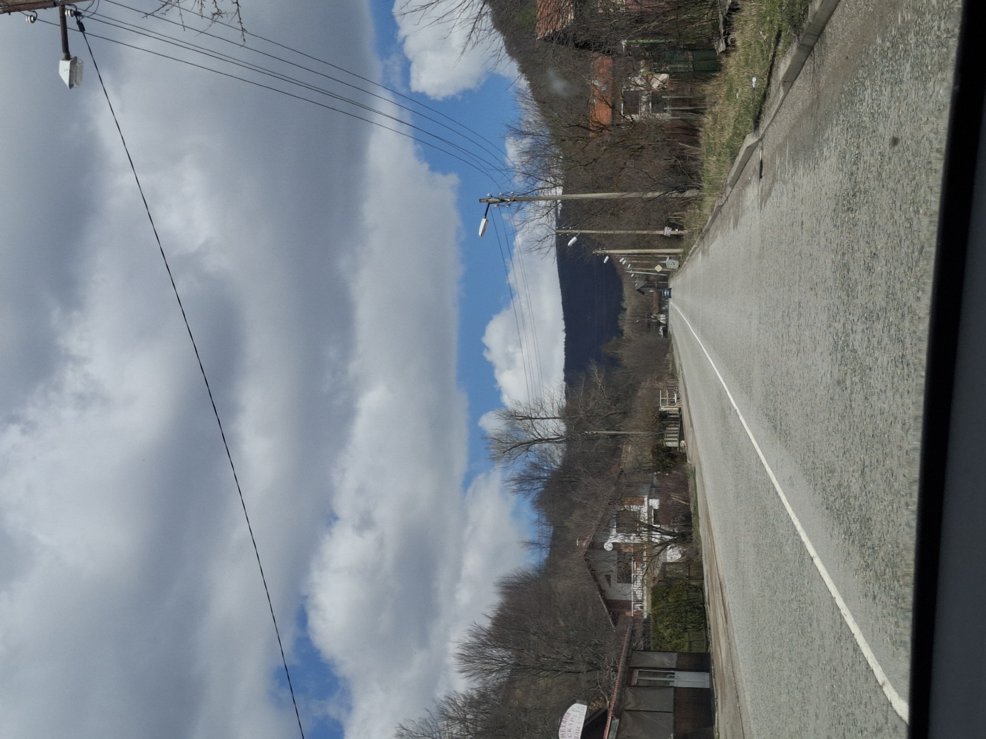 The image depicts a road in Gintsi, Bulgaria. It's a straight, paved road with a white dividing line. Utility poles with wires run parallel to the road. Buildings and trees line the roadside. A mountain is visible in the distance. The sky is partially cloudy.