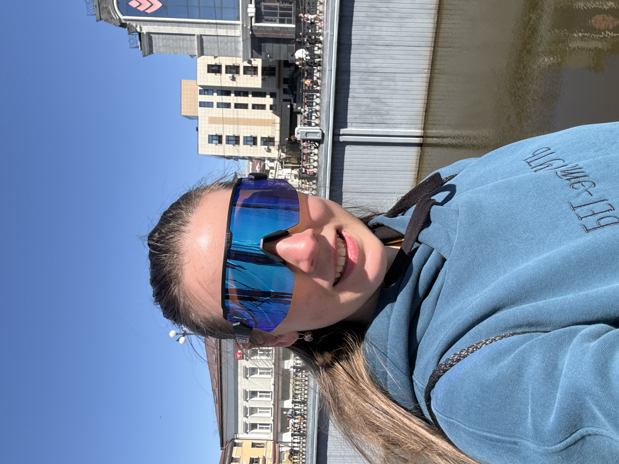 A selfie image shows a light-skinned person with long brown hair wearing blue sunglasses and a blue hoodie. The location appears to be Kazan, Russia, based on the cityscape in the background. The subject is on a bridge or walkway with a body of water behind a gray metal railing. Buildings of varying architectural styles are visible in the background, along with people walking along the waterfront. The sky is clear and blue.