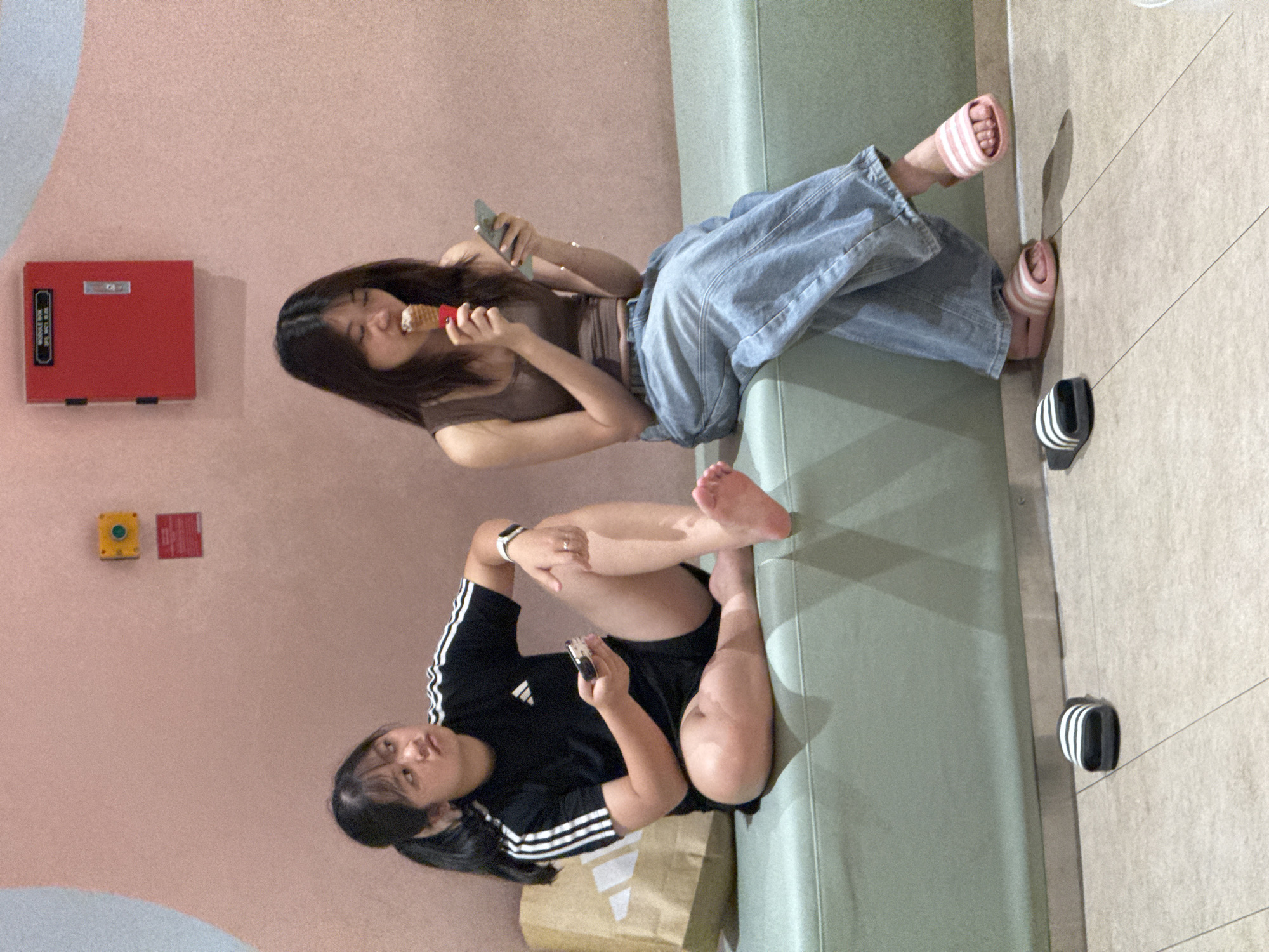 Two young women are seated on a light green bench in what appears to be an indoor waiting area, possibly in Hanoi, Vietnam. The wall behind them is a soft pink with a curved light blue section on the left and a red fire alarm box mounted on the upper right.
The woman on the left, wearing a black Adidas t-shirt and black shorts, is looking to her left with a slightly confused or questioning expression. She is holding a mobile phone in her right hand and appears to have a ring on her left ring finger. Her legs are crossed, and her bare feet are visible. Next to her is a brown paper shopping bag with the Adidas logo.
The woman on the right is wearing a brown tank top and light blue baggy jeans. She is eating an ice cream cone and looking down at her mobile phone, which she holds in her left hand. She is wearing pink and white striped slide sandals.
On the floor in front of the bench are two pairs of slide sandals: black and white Adidas slides in front of the woman on the left, and pink and white slides in front of the woman on the right. The floor is tiled in a light neutral color.