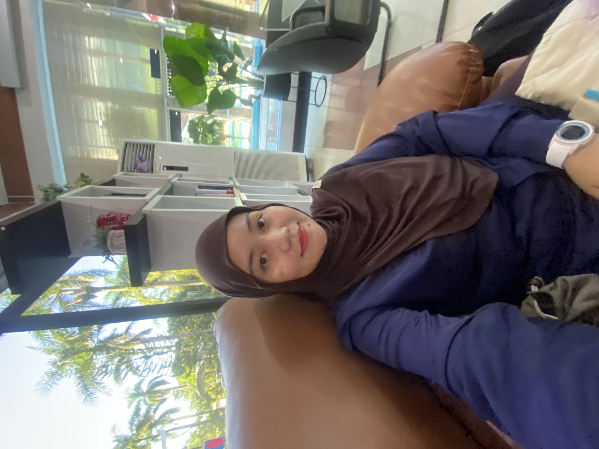 A young woman wearing a brown hijab and a navy blue long-sleeved top is seated on a brown leather sofa, looking towards the camera. Her right arm is extended, likely holding the device used to take the selfie. On her left wrist, she wears a white smartwatch with a blue bezel. The background shows an indoor setting with white shelving units containing books and decorative items. Beyond the shelves, through a large window, palm trees and a clear sky are visible, suggesting a tropical or subtropical environment. The location is the city of Banjarbaru, Indonesia.