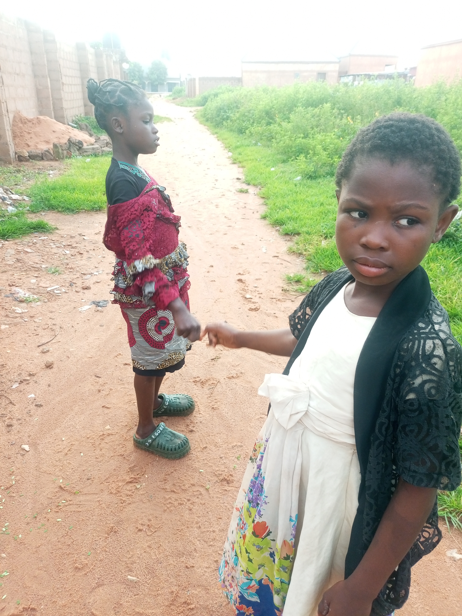 Two young girls stand on a dirt road in Tempure, Nigeria. The girl in the foreground, on the right, wears a white dress with a floral print, a black lacy cardigan, and appears to be looking off to the side with a serious expression. The girl in the background, on the left, faces away from the camera, wearing a dark top with a patterned maroon overlay and green clogs. The road is lined with dry grass and sparse vegetation, with a low wall made of concrete blocks visible on the left side of the frame. The sky is overcast, creating a soft, diffused light.