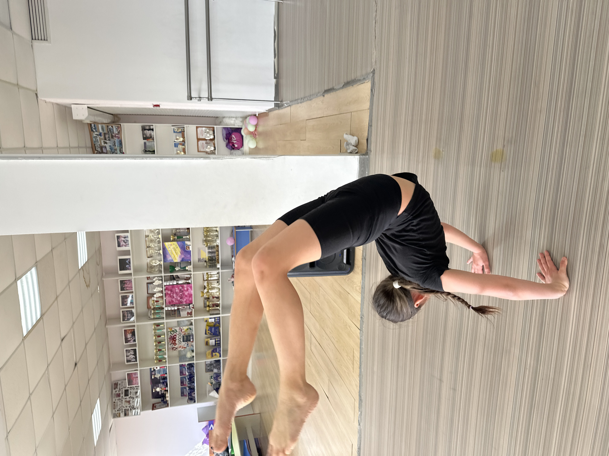 The image depicts a young person performing a backbend, also known as a bridge pose, in an indoor studio, likely located in Votkinsk, Russia. The individual is dressed in black athletic wear, consisting of a t-shirt and shorts, with their dark hair tied back in a braid. They are barefoot, with their hands and feet planted on the light-colored wooden or laminate floor, arching their body upwards.
The setting appears to be a dedicated training or practice space, possibly a dance studio or gymnastics gym. The background features extensive shelving units filled with numerous trophies, awards, and framed photographs, indicating a history of accomplishments. Among these awards, some small, light-colored balloons (pink, white, light green) are visible. The walls are white, and the ceiling is a tiled drop ceiling with several rectangular fluorescent light panels providing bright illumination.
While most text on the trophies and awards is unreadable due to distance and blur, some Cyrillic characters are faintly visible, such as "ИПЦ" and "ПРИ" on different awards. The overall impression is one of an athletic environment focused on physical skill and achievement.