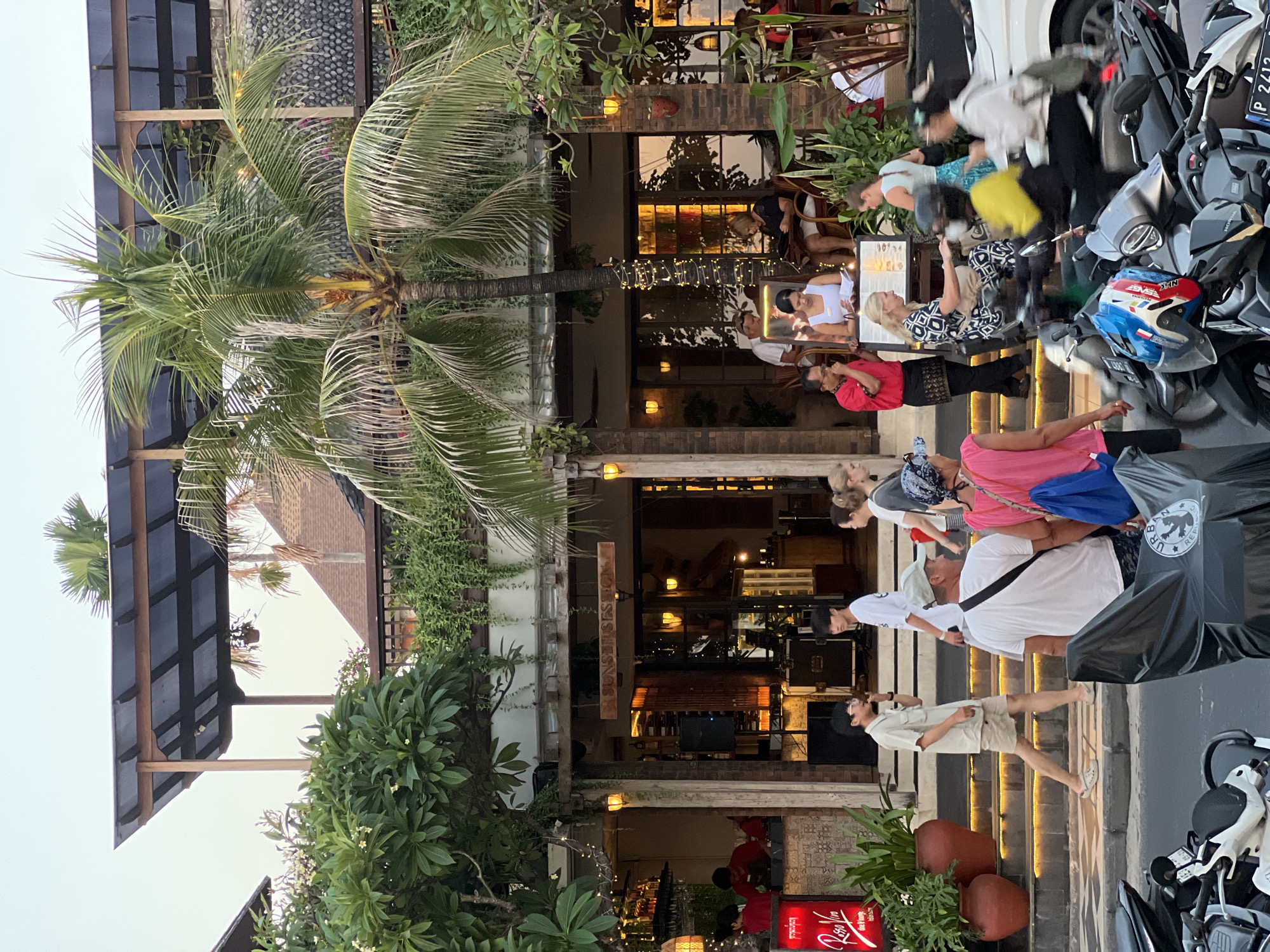 In Kuta, Indonesia, a street scene unfolds outside a multi-level establishment with an outdoor dining area and upper-level balcony. Palm trees and lush green foliage frame the building, which features wooden beams, a translucent roof, and hanging string lights. People are depicted walking on the street and sidewalk, entering and exiting the building, and seated at tables. A group of individuals is gathered near the entrance, while others are in the process of ordering or dining. Motorbikes are parked along the street, with some individuals on or near them. The architecture suggests a tropical, possibly resort-style atmosphere.