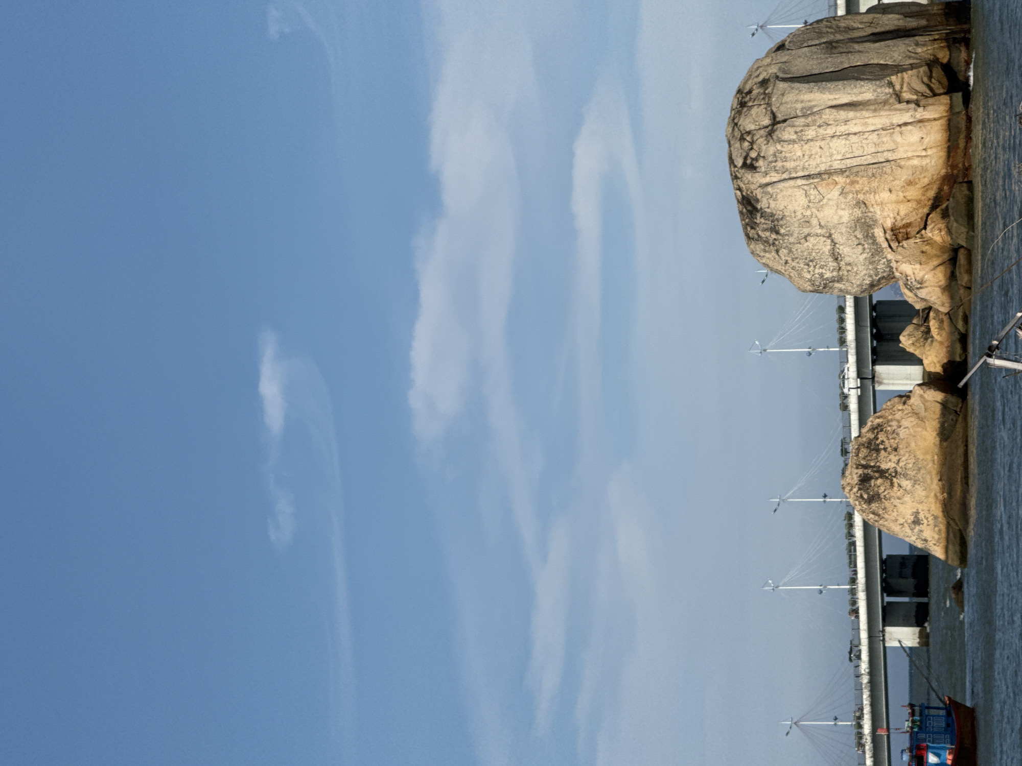 The image depicts a coastal scene in Bac Nha Trang Ward, Vietnam. In the foreground, a large, weathered rock formation dominates the right side of the frame, with smaller rocks visible around its base. The water is a deep blue with moderate waves. A small fishing boat, painted blue and red, is positioned on the left side of the image, partially submerged in the water. In the mid-ground, a long, modern bridge stretches across the water, supported by pillars and featuring suspended cables. Vehicles are visible on the bridge, suggesting it is an active transportation route. The sky is a clear, pale blue with a few wispy clouds. The overall impression is one of a calm, scenic day with a blend of natural and man-made elements.