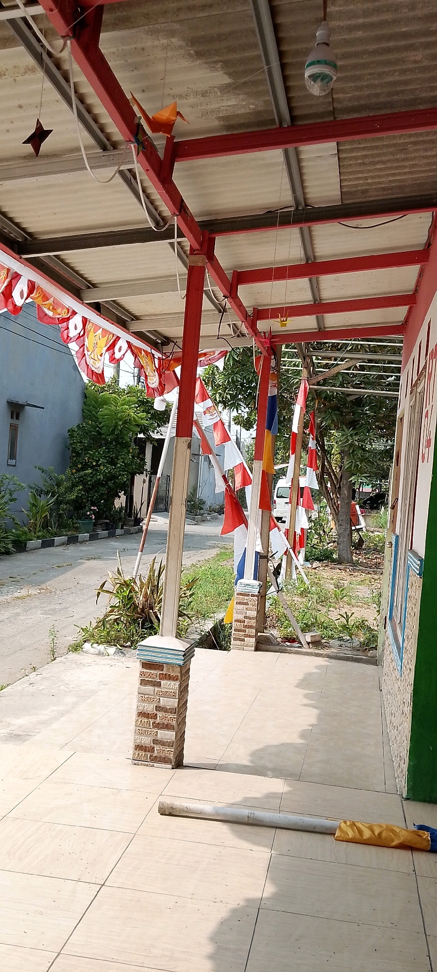 The image depicts an outdoor walkway or patio area under a red-framed roof structure. Red and white Indonesian flags are strung along the roofline and also hang vertically from poles. A light fixture is visible hanging from the corrugated ceiling. On the left side of the frame, a light blue building with a window is visible, adjacent to lush green foliage. The walkway is paved with large, light-colored square tiles. In the foreground, a long, cylindrical object, possibly a broom or mop handle, lies on the tiles with a yellow cloth object attached to one end. Brick pillars support the roof structure. The environment suggests a residential or public space, possibly decorated for a national celebration, given the prominent display of Indonesian flags. The background includes a paved road and more greenery.