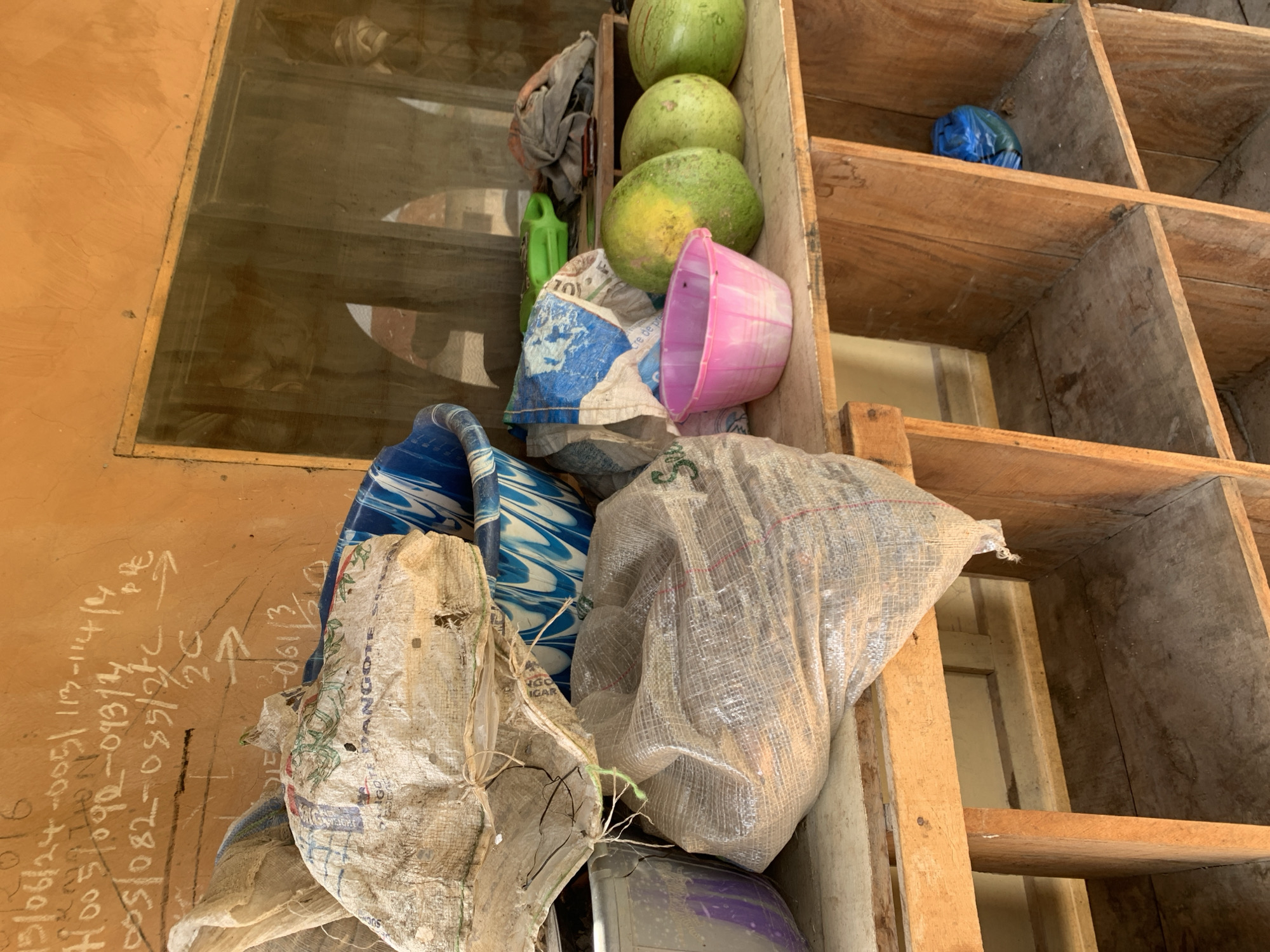 The image shows a close-up view of a wooden shelving unit filled with various items, primarily bags and some green fruits that appear to be watermelons. The shelves are dusty and weathered, suggesting an outdoor or storage setting. In the foreground, several sacks are piled up, one is made of white and blue woven material, another is a larger, translucent sack filled with what looks like soil or sand. A pink plastic container is also visible.
To the left of the shelves, a wall with chalk markings is partially visible. The markings include numbers and dates, some in English and some in a local language, suggesting some form of inventory or record-keeping. The presence of the fruits and sacks implies that this might be a storage area for produce or goods. The lighting suggests it is daytime, but there are no clear indicators of the weather. The overall impression is of a functional, perhaps rural or market-related storage space in Jos, Nigeria. No people are visible in the image.