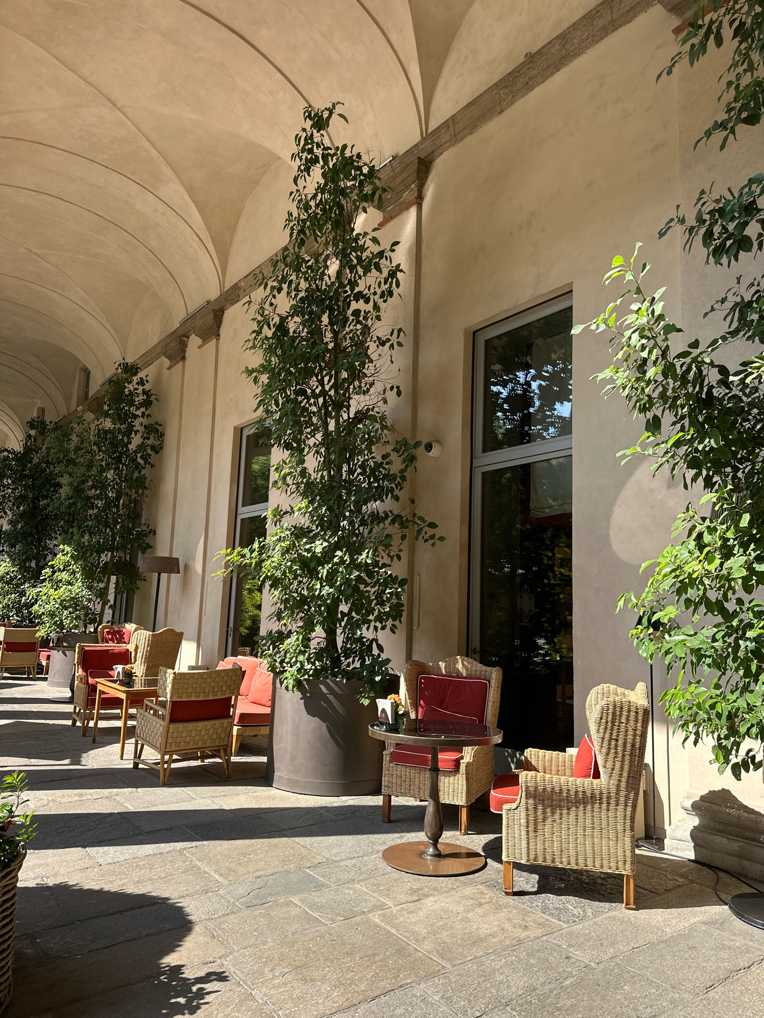 The image shows an outdoor seating area of a building with an arched ceiling in Milan, Italy. The seating area is paved with large stone tiles and features several tables and chairs made of wicker. The chairs are furnished with red cushions. Tall potted plants are placed around the seating area, and the building itself is light-colored with large windows. Sunlight casts shadows on the ground, indicating a sunny day. In the background, part of the arched walkway is visible, and more potted plants line the wall. The scene appears to be a tranquil outdoor space, likely a cafe or restaurant patio.