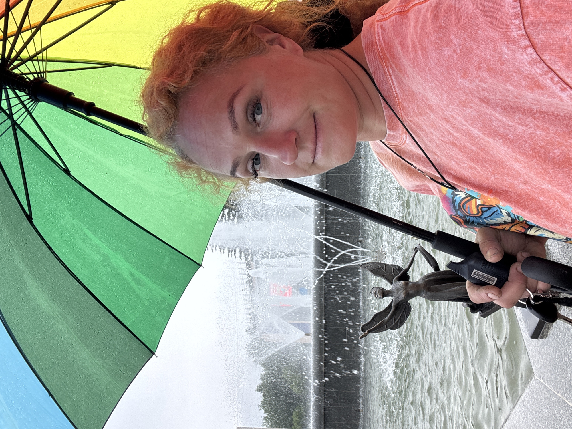 A woman with curly blonde hair and blue eyes is holding a rainbow-colored umbrella while standing near a fountain. The umbrella is open, and water droplets are visible on its surface, suggesting it is raining. The woman is wearing a pink t-shirt. In the background, a fountain is spraying water, and a bronze statue of a winged figure is visible in the water. The scene appears to be outdoors in a public space. There is no indication of this image being taken in Chaikovsky, Russia.