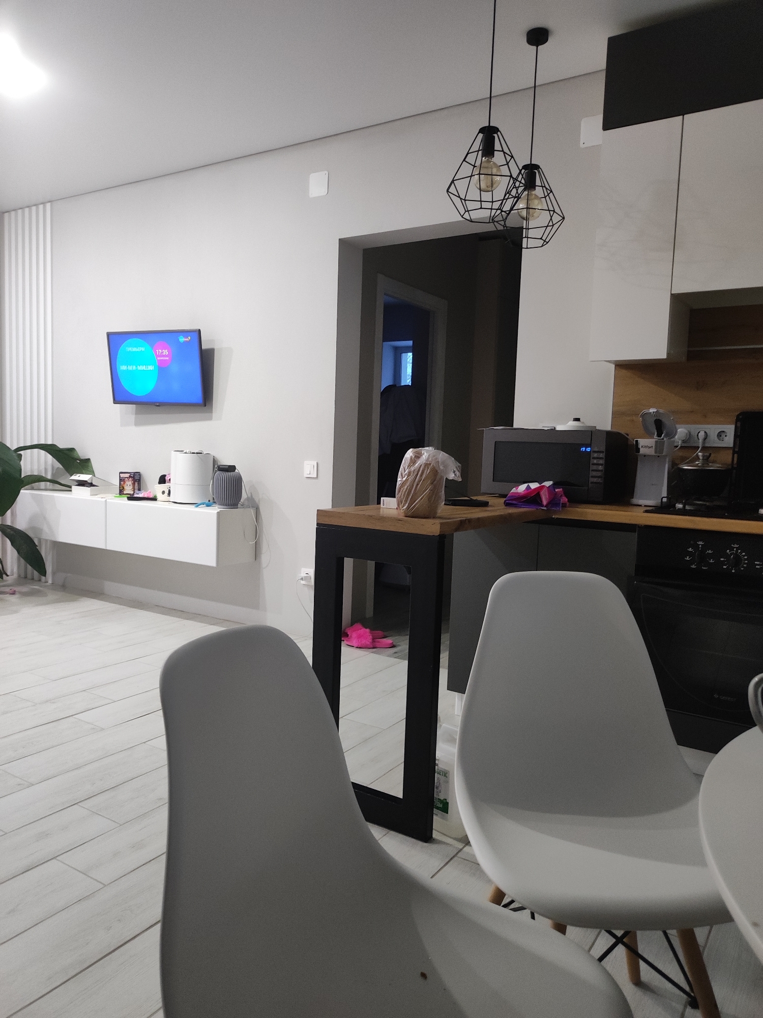 The image depicts an interior scene, likely a room within an apartment, possibly located in Votkinsk, Russia based on the display on the TV screen. The room features a living area transitioning into a kitchen. The living area has a white floating shelf unit, upon which are various decorative objects including a humidifier. Above the shelf is a flat-screen TV. A large green plant sits near the unit.
In the kitchen area, there's a counter with a black and wood bar table extension. Atop the counter are kitchen appliances, including a microwave and a coffee maker. Food is present, along with pink slippers on the floor. Above the counter are white kitchen cabinets with dark upper cabinets. Two modern, geometric pendant lights hang above the bar area.
In the foreground, two white dining chairs are partially visible, suggesting a dining area. The walls are painted a neutral gray. The floor is covered with light-colored, wood-look planks. A doorway is visible leading to another room, revealing a glimpse of a light blue wall. The time displayed on the microwave is 19:10, while the TV displays 17:35.