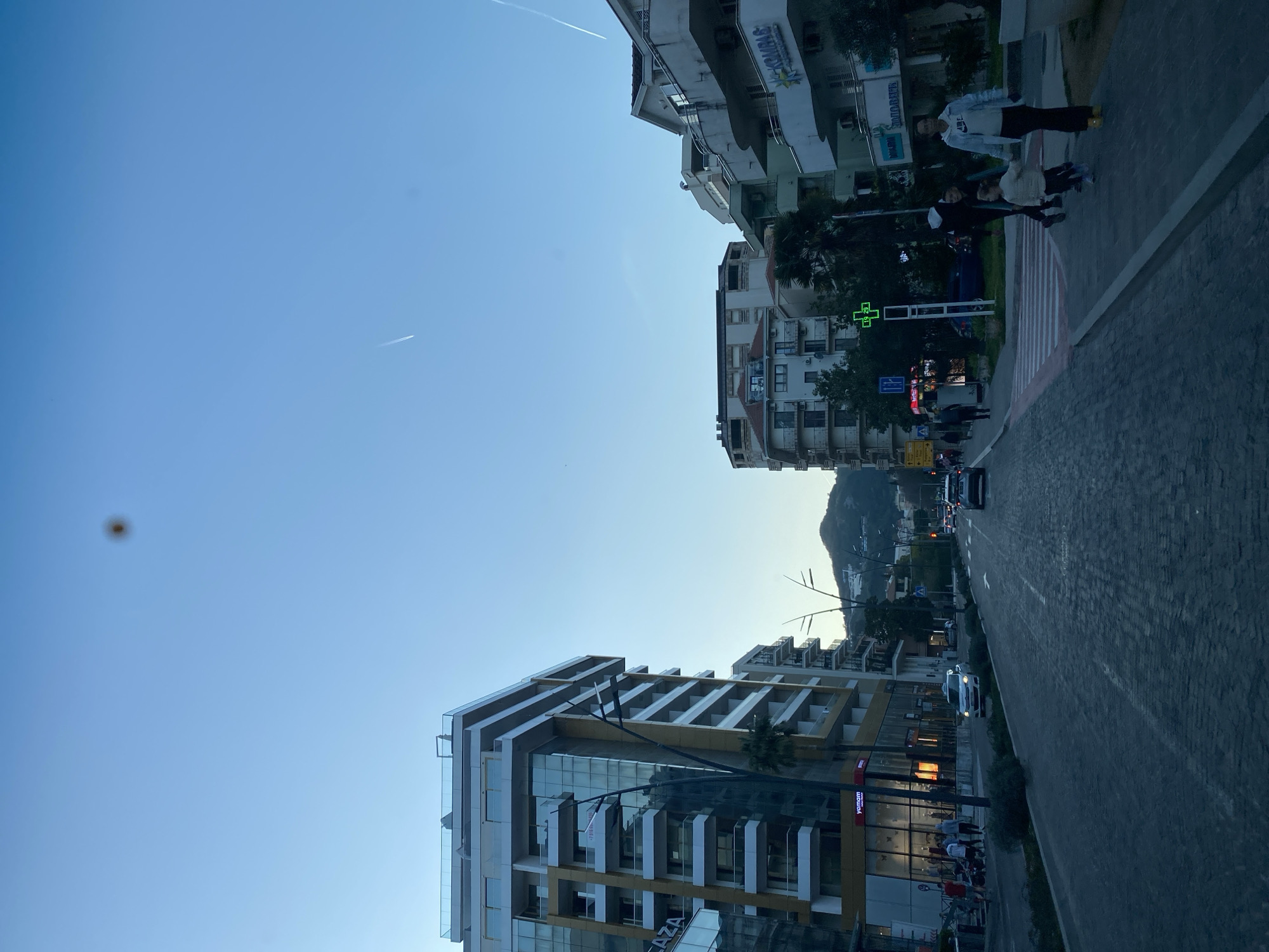 The image depicts a street scene in Budva, Montenegro, with buildings on either side of a road. The sky is blue. Two contrails are visible. On the right, people are walking along a sidewalk. A pharmacy is indicated with a green cross. Traffic moves down the street. In the background, a mountain is visible. Modern buildings line the left side of the image.
