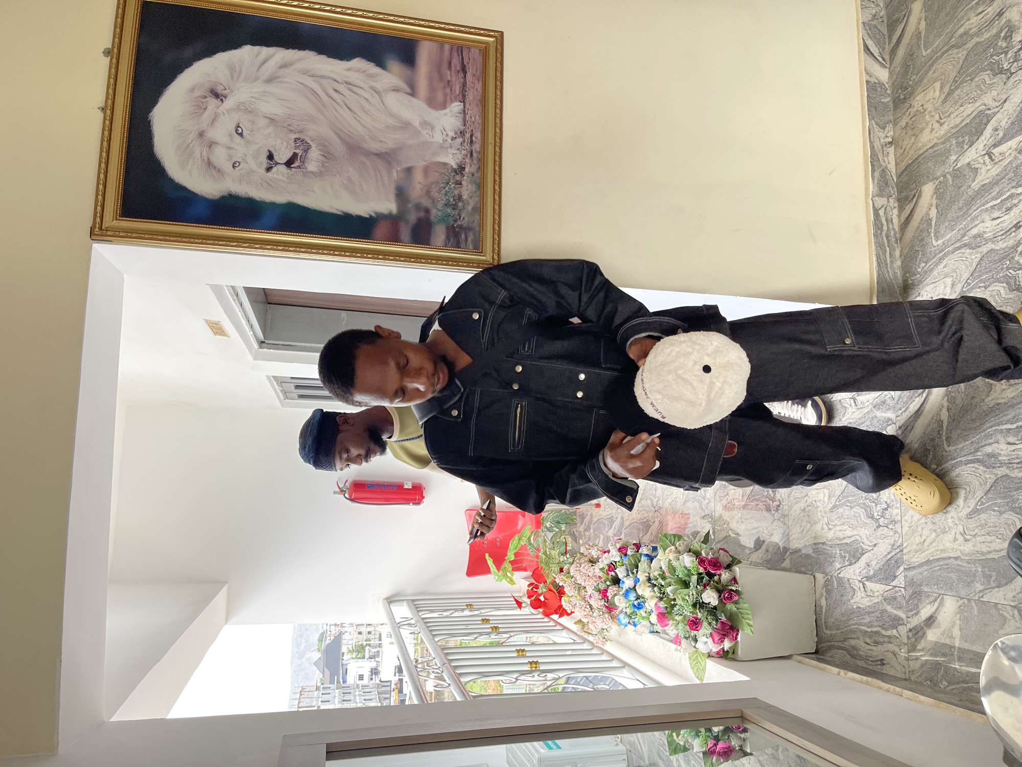 The image depicts an indoor scene, likely a hallway or entrance area in Gwarinpa, Nigeria, featuring two men and various decorative and functional objects. The main subject, positioned centrally, is a man wearing a dark denim jacket and matching pants with contrasting white stitching. He wears light-colored, possibly foam, slip-on shoes. In his left hand, he holds a round, fluffy white object with the text "Alexander Wang" visible on it, while his right hand holds what appears to be a pen or marker.
Behind him, partially obscured, is another man wearing a dark cap. The walls are a mix of light yellow and white. High on the left wall hangs a large, ornate golden-framed painting of a majestic white lion with piercing blue eyes against a dark background. Further back, a red fire extinguisher is mounted on a white wall.
The floor is covered with grey and white marble-patterned tiles. A decorative metal railing with a floral arrangement of artificial pink, blue, white, and red flowers in a white planter box separates the interior from an outdoor view. Through the railing, buildings and distant hills are visible under a bright sky, indicating it is daytime. The scene suggests a well-lit, modern indoor space with a view of an urban landscape.