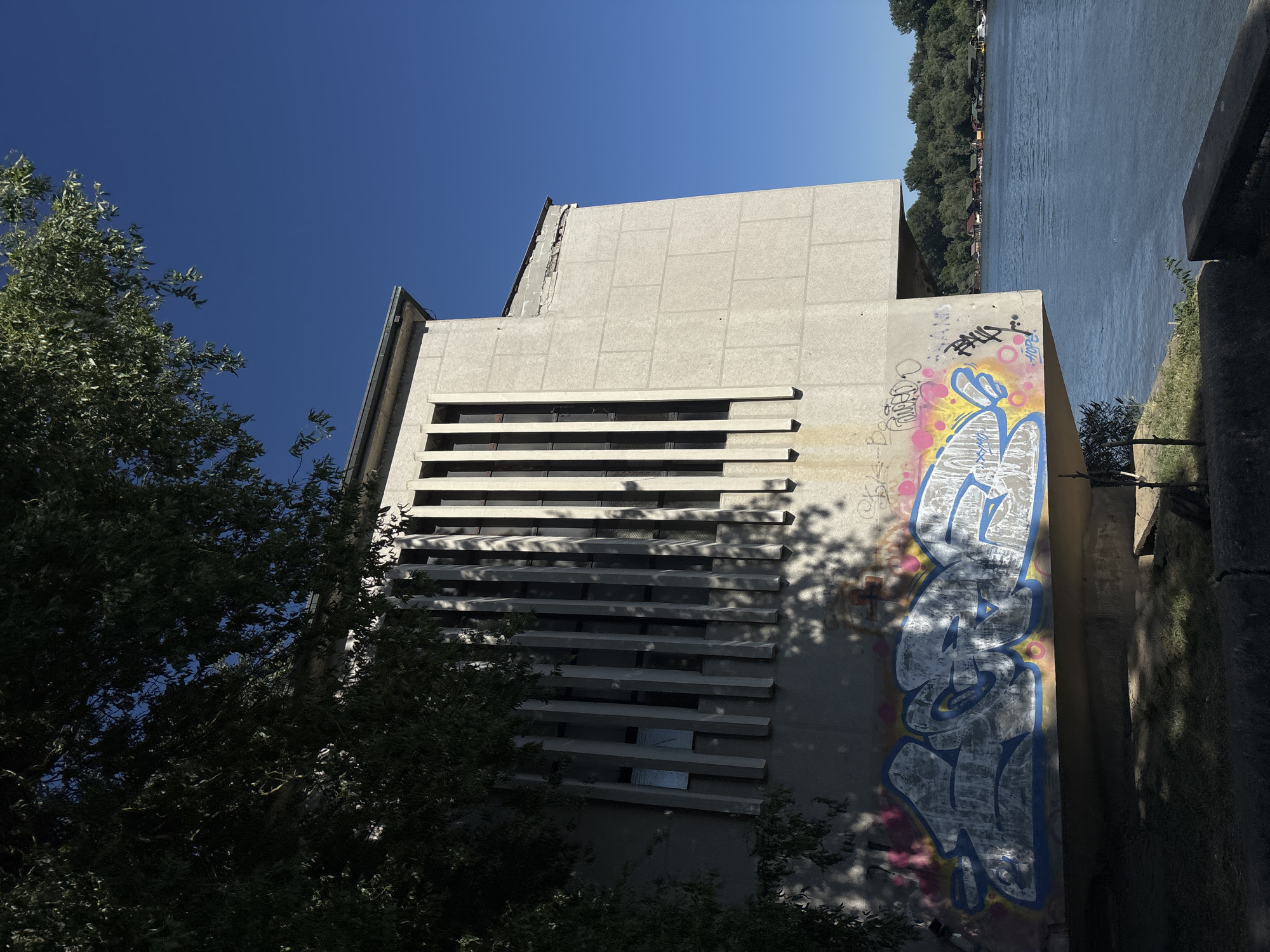 The image captures a concrete building in Belgrade, Serbia. The structure is characterized by a repetitive pattern of horizontal concrete beams interspersed with rectangular windows. Graffiti is visible on the building's exterior, adding an element of urban decay. A mature tree with dense foliage partially obscures the lower left portion of the building, casting shadows on the wall. Adjacent to the building, a body of water is visible, suggesting a riverside location. The sky is clear and blue, providing a backdrop that contrasts with the grey concrete. The perspective of the photograph is from a slightly low angle, enhancing the height of the building. The presence of graffiti and the overall architectural style suggest an industrial or institutional building in a potentially transitional area of the city. The image doesn't show people.