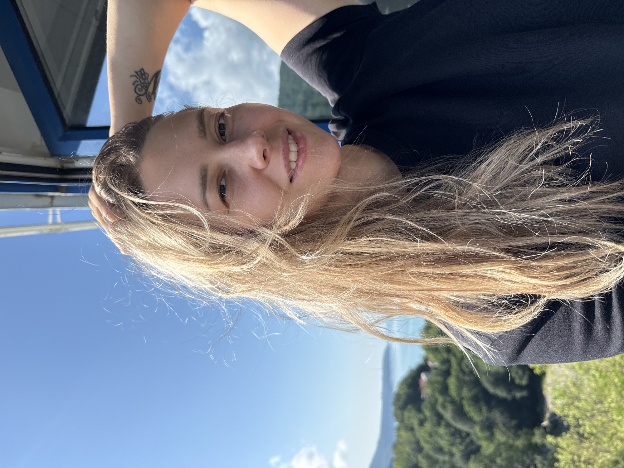 A woman with blonde hair and a tattoo on her arm is taking a selfie outdoors. She is wearing a black t-shirt. In the background, there is a body of water, lush green trees, and a bright blue sky with some clouds. The scene appears to be a scenic overlook in or near the city of Nebug, Russia, possibly from a cable car as suggested by the blue frame in the upper right corner. The lighting suggests it is daytime and sunny.