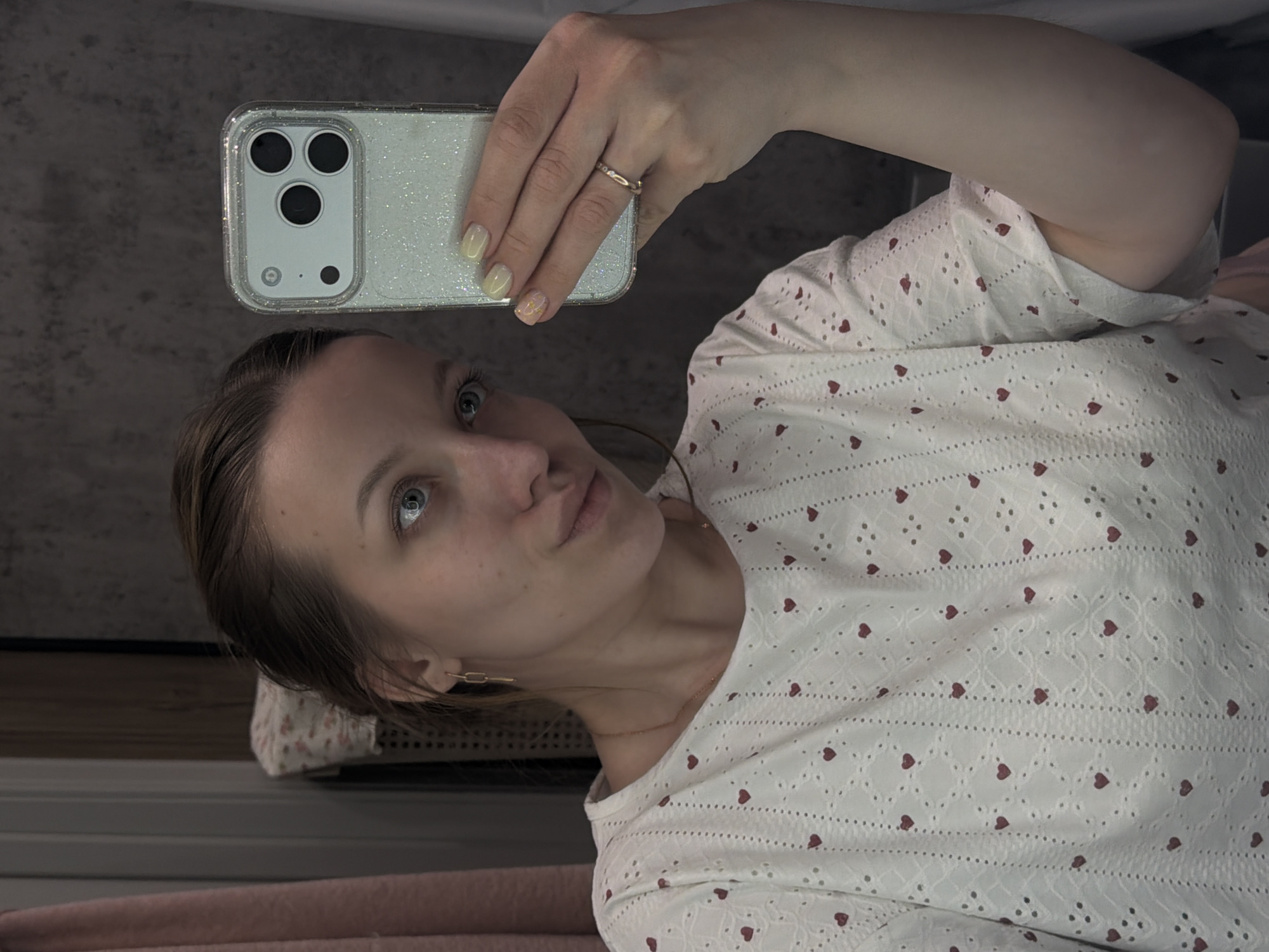 A woman with light skin and blue eyes is taking a selfie with a smartphone. The phone has a white, glittery case and three visible camera lenses. The woman is wearing a white top with small red heart patterns. A gold ring is visible on her left ring finger. She has gold earrings and a gold necklace. The background appears to be an indoor setting with a wooden surface visible. The location in Reshetnikovo, Russia is not discernible from this image.