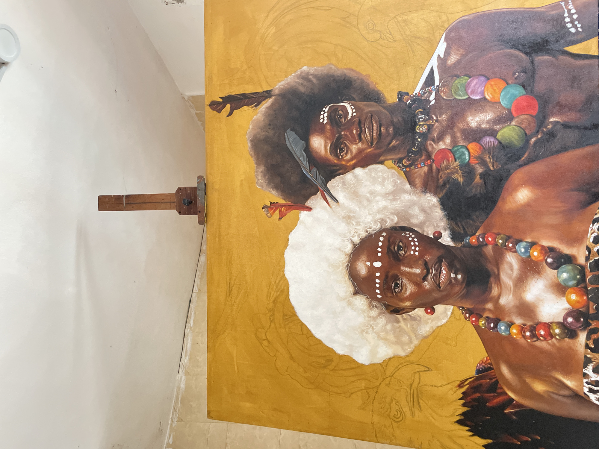 The image depicts a painting in progress. Two dark-skinned figures with tribal markings and colorful beaded necklaces are the central subjects. One figure has a large white fluffy headdress, while the other has a dark Afro with feathers. The background is a gold color with faint sketches of other elements yet to be painted. The painting is on an easel in a room with white walls. The location is likely Ado, Nigeria.