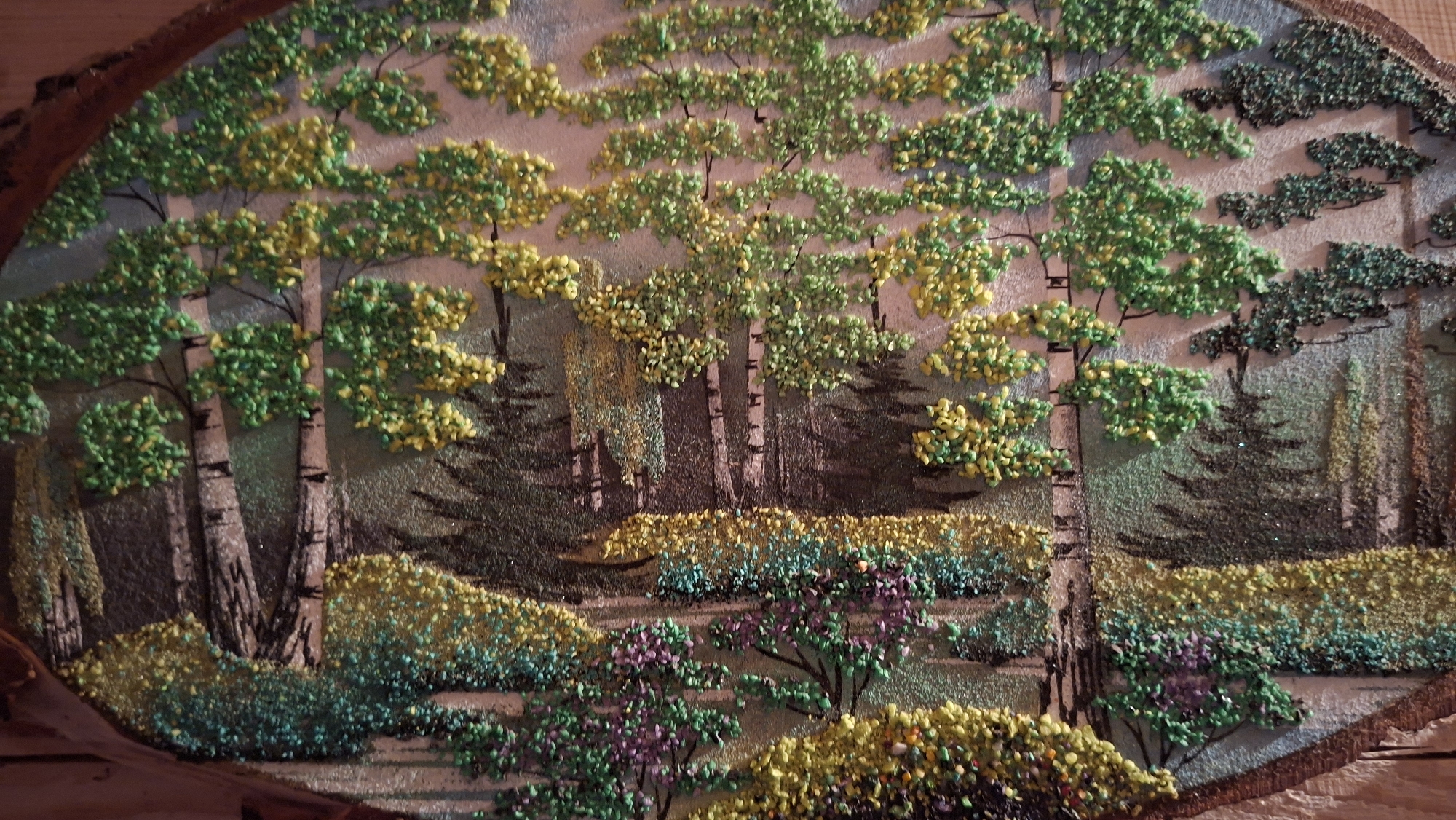 The image shows a landscape artwork depicting a forest scene, likely a decorative piece or craft project. The scene includes various trees, predominantly birch trees with their characteristic white bark and dark markings, along with other evergreen trees in the background. The foliage is represented with green and yellow hues, possibly suggesting different seasons or varieties of trees. In the foreground, there are bushes and shrubs, with a mix of green, yellow, and hints of purple, adding depth and color to the composition. The artwork appears to be textured, possibly created using small stones, beads, or paint applied in a pointillistic style. The background has a muted gray-brown tone, possibly representing the sky or distant landscape. The artwork is framed by a rough, natural border, suggesting it is mounted on a wooden base. There is no evidence that it is located in Большой Букор, Russia.