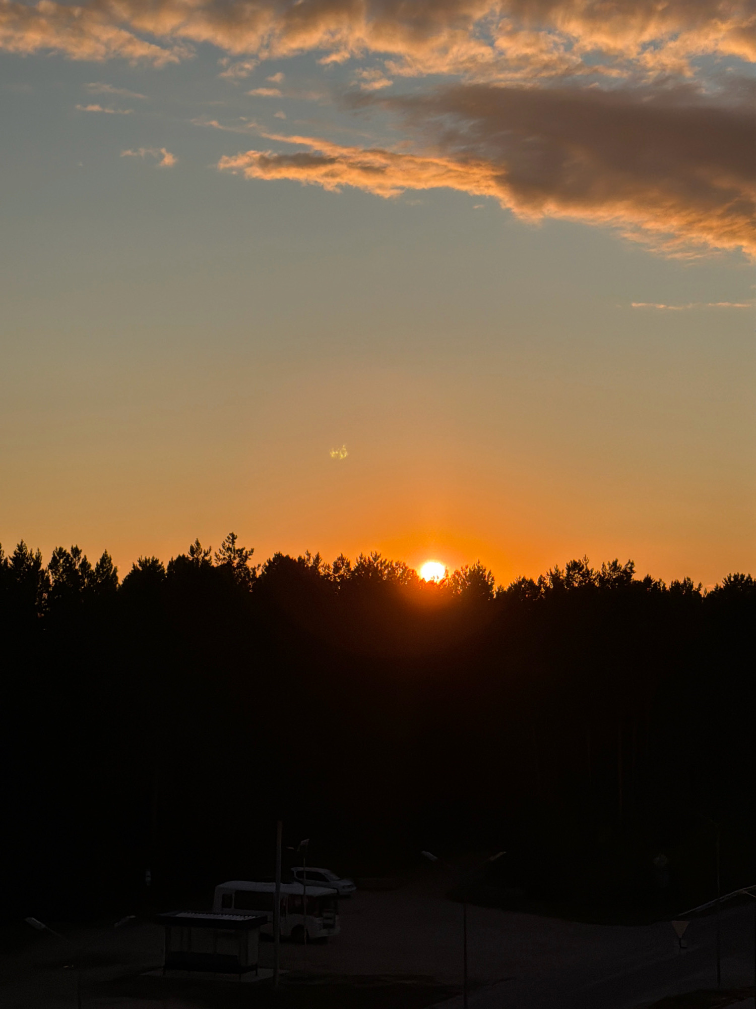 The image depicts a sunset over a silhouetted forest in the city of Tsiolkovsky, Russia. The sky transitions from a pale blue near the top to a vibrant orange near the horizon, with streaks of pink and yellow clouds. The sun, partially obscured by the treeline, casts a warm glow. In the foreground, a dark, flat area suggests a road or open space. Visible are a bus stop shelter with a dark roof and bench, and two vehicles. One is a white van or minibus parked behind the bus stop, and a second, lighter-colored car is parked further in the background. Streetlights are also present, with one clearly visible near the bus stop and another further back. The overall scene is tranquil and captures the end of daylight.