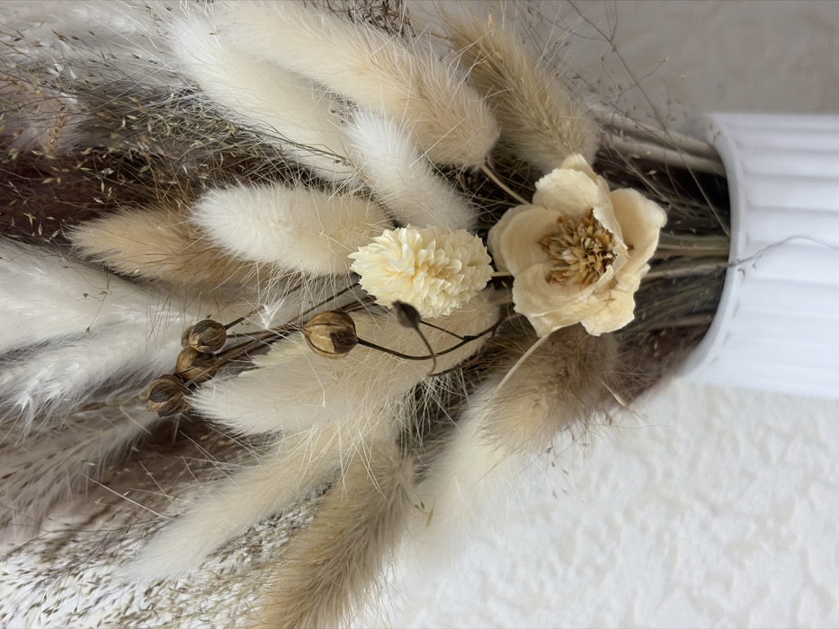 The image displays a close-up shot of a decorative arrangement primarily composed of dried flowers and grasses, presented in neutral, earthy tones. The arrangement is dominated by fluffy, elongated seed heads, likely Lagurus ovatus (bunny tail grass), in varying shades of cream, off-white, and light beige. These are interspersed with thinner, wispy strands of dried grasses in brownish and grayish hues, adding texture and depth to the bouquet.
At the center-right of the arrangement, there are two distinct dried flowers. The larger one is an open bloom, delicate and light cream in color, with visible stamens or a central cluster of petals in a slightly darker beige. To its left, a smaller, spherical flower with a tightly clustered, intricate petal structure is visible, in a pale yellow or off-white shade. Below and to the left of these flowers, a cluster of small, round, dried seed pods or berries, dark brown in color, are attached to thin, wiry stems.
The overall color palette is very subdued, consisting mainly of whites, creams, beiges, light browns, and grays, contributing to a rustic and natural aesthetic. The textures are diverse, ranging from the soft, feathery quality of the fluffy grasses to the delicate dryness of the flowers and the smooth, hard appearance of the seed pods.
On the far right side of the image, a portion of a white, vertically ribbed object is visible, suggesting it might be a vase or a decorative container holding the arrangement. The background is softly lit and appears to be a light-colored, possibly textured wall or surface, further emphasizing the foreground elements. The lighting is soft and even, highlighting the intricate details and textures of the dried botanical elements.