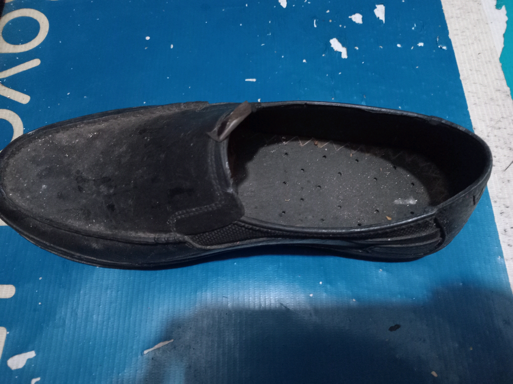 A single black shoe is pictured lying on a textured blue surface. The shoe is a slip-on style, and the interior of the shoe is visible, showing a perforated insole. The shoe appears to be made of synthetic material and has a low heel. The blue surface has some white specks and a white stripe running vertically on the right side of the image. The lighting suggests an indoor setting, possibly a workshop or storage area, given the utilitarian nature of the surface and the shoe. There are no people or other objects clearly visible in the frame. The location is Unknown, Unknown.