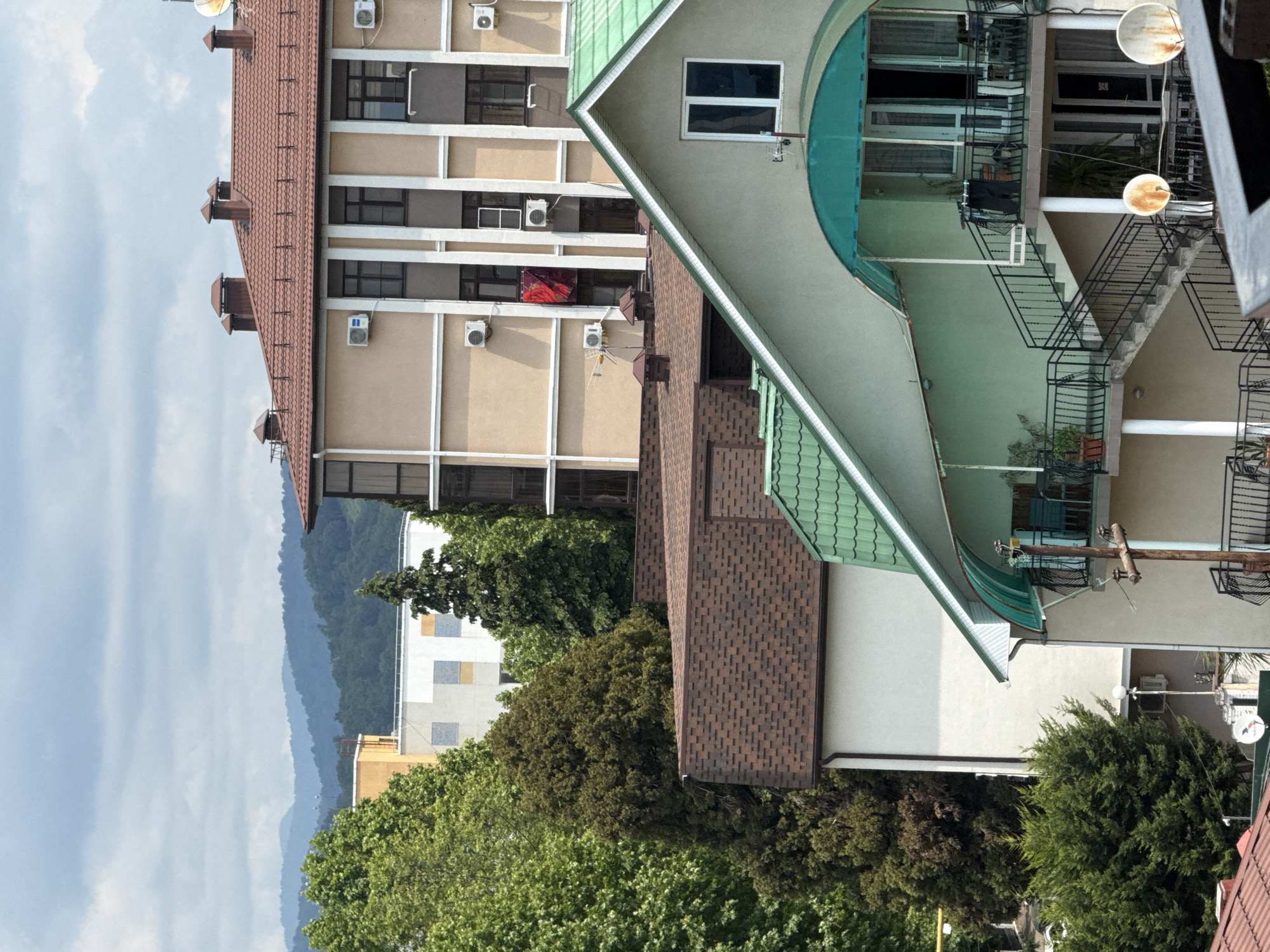 The image depicts a collection of multi-story buildings and lush greenery, suggesting a dense urban environment with a significant presence of trees. The buildings have a variety of architectural styles and roof types, including a prominent brown tiled roof and a green tiled roof. Several windows and balconies are visible, some of which are adorned with plants. Air conditioning units are mounted on the exterior walls of one of the buildings. A red cloth hangs from a window of a light-colored, multi-story building. In the background, a series of rolling hills or mountains are visible, partially obscured by trees and clouds. The sky is overcast with a light, diffused quality of light. There are no people visible in the frame. The overall impression is one of a residential or commercial area with a temperate climate, likely situated in or near a city.
Given the context that this image is from Sochi, Russia, the scene is consistent with the city's geography, which is known for its mix of urban development and proximity to mountainous terrain. The architecture of the buildings, the abundant vegetation, and the cloudy sky are all characteristic of the Black Sea coast region. The presence of multiple buildings packed closely together, with some indicating commercial or hospitality use (e.g., balconies, multiple stories), could suggest an area within the city or its immediate surroundings where tourism or residential development is prevalent. The overall setting is a complex urban landscape intertwined with natural elements, typical of coastal cities in mountainous regions.