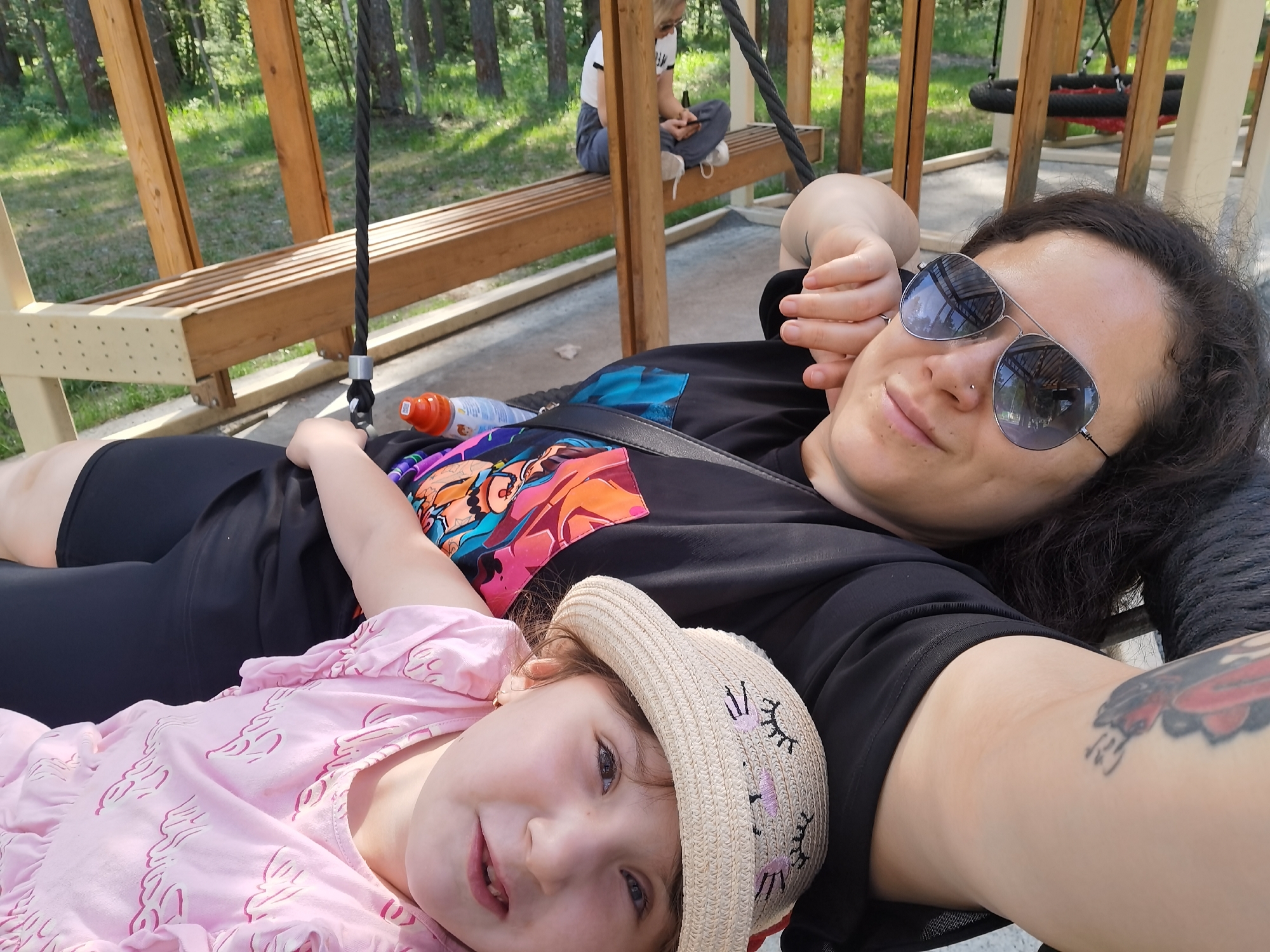 The photograph shows a woman lying down with a young child in her arms. They are on a swing within a playground, as evidenced by the wooden beams, benches, and other playground equipment visible in the background. The woman is wearing sunglasses and a black t-shirt, while the child is wearing a pink dress and a straw hat with embroidered eyelashes. A person is sitting on a bench in the background. The setting appears to be in a park or recreation area in Perm, Russia, as the metadata indicates the location.