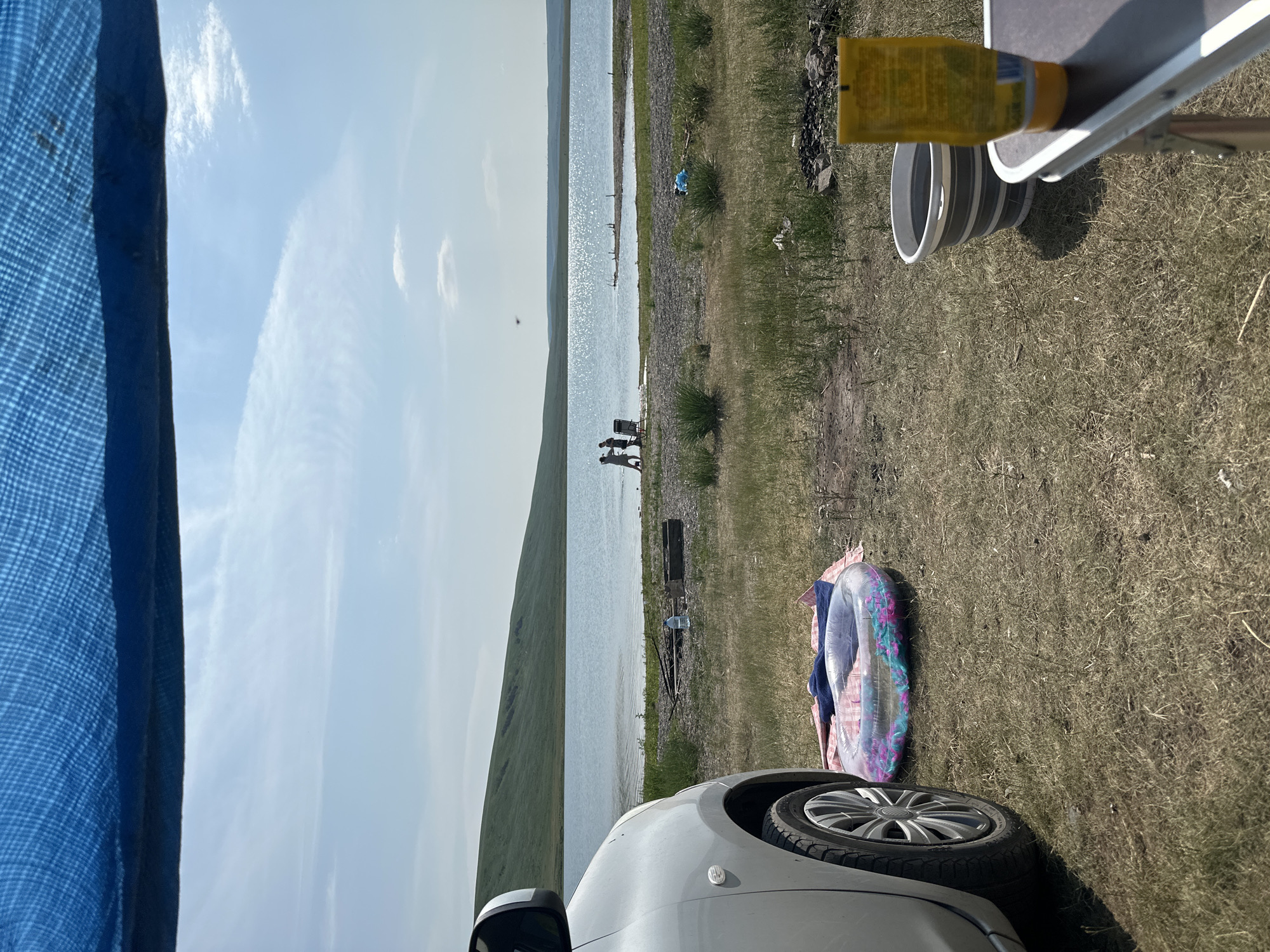 This image captures a serene daytime scene by a body of water, likely a lake or reservoir, in Novonikolaevka, Russia. The sky is a bright blue with wispy white clouds, suggesting fair weather. In the foreground, the edge of a silver car is visible, with its wheel resting on dry, grassy ground. Beside the car, a colorful inflatable ring and a pink blanket are laid out, hinting at recreational activities like swimming or sunbathing.
Further in the distance, a gentle, green hillside slopes down towards the water's edge. Along the shoreline, several figures, appearing to be people, are standing and interacting near what might be a chair. The water itself is calm and reflects the bright sky, with some areas sparkling under the sunlight. A few small wooden posts or remnants of structures are visible in the water.
To the right side of the frame, a portable table is set up, with a yellow bottle of what appears to be sunscreen or lotion resting on it. A grey, collapsible bucket is also present. The ground around the table and to the right is covered in dry grass and scattered small rocks. The overall mood is relaxed and peaceful, indicative of an outdoor leisure activity on a pleasant day. The scene is bathed in daylight, likely mid-morning or afternoon.
