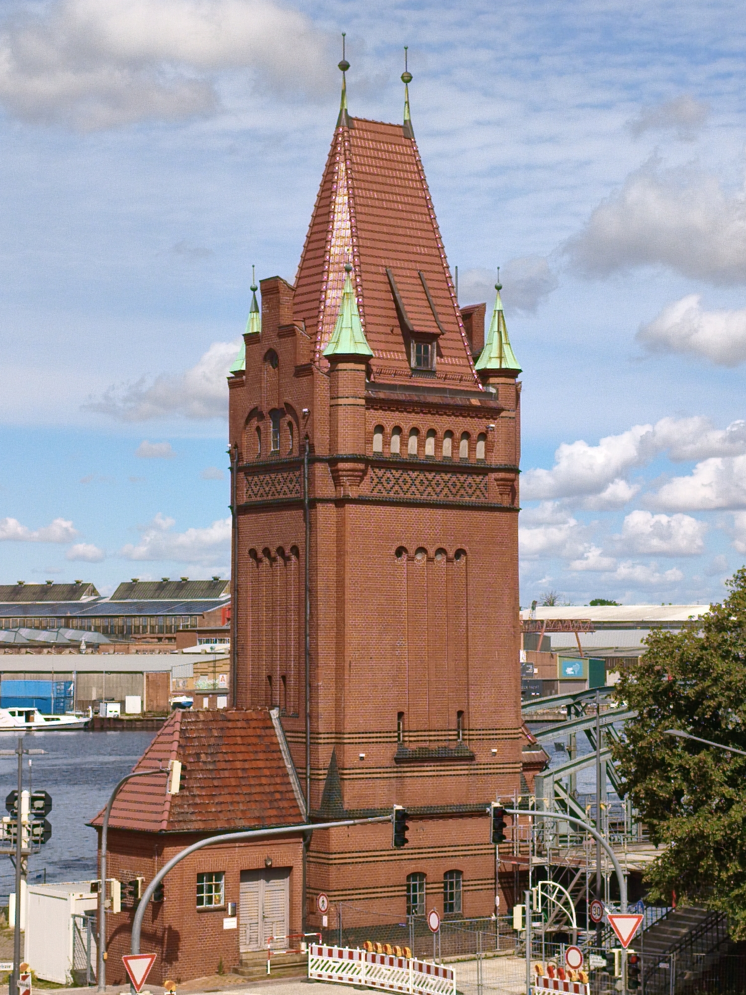This image shows a view of the historic Holstentor in Lübeck, Germany, a UNESCO World Heritage site. The Holstentor is a 15th-century brick gatehouse that served as a city gate and a defensive structure. It is a prominent landmark in Lübeck and a symbol of the city's Hanseatic past.
The image captures the Holstentor from a slightly elevated perspective, with the iconic twin towers and red brick façade clearly visible. The sky above is a vibrant blue with fluffy white clouds, suggesting fair weather. The Holstentor is situated near the Trave River, and a portion of the river is visible in the foreground, with a boat docked nearby.
The surroundings of the Holstentor are a mix of historic architecture and modern elements. In the background, there are other buildings and structures, some of which appear to be industrial or port-related, reflecting Lübeck's history as a major trading port. There are also some signs of ongoing construction or renovation, with scaffolding and barriers visible in some areas.
The image appears to be taken during the daytime, with the sun casting shadows and highlighting the details of the architecture. The overall impression is one of historical significance and enduring presence, with the Holstentor standing as a testament to Lübeck's rich past.