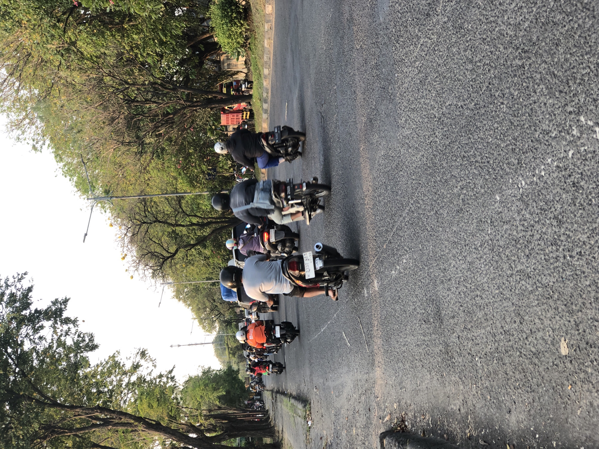 The image depicts a street scene in Gelam, Indonesia, with several motorcycles carrying riders and passengers proceeding along a paved road. The road is lined with trees on the left and right, with some buildings and streetlights visible in the background. The sky is bright, indicating it is daytime. A variety of motorcycles are in motion, with riders wearing helmets and casual clothing. Some motorcycles are carrying single riders, while others have passengers. Traffic appears to be moderate, with no apparent congestion. The overall impression is of a typical urban street scene in Indonesia during daylight hours.
