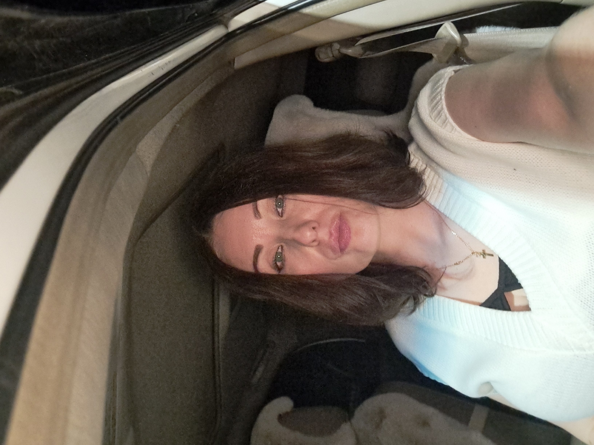 A woman with dark brown hair and green eyes is seated in the passenger seat of a car. She is wearing a white knitted sweater over a black top and a gold cross necklace. The interior of the car is visible, including the headrests of the front seats and the ceiling lining. The image appears to be taken at night, as indicated by the lighting and the darkness outside the car. The location is not specified.
