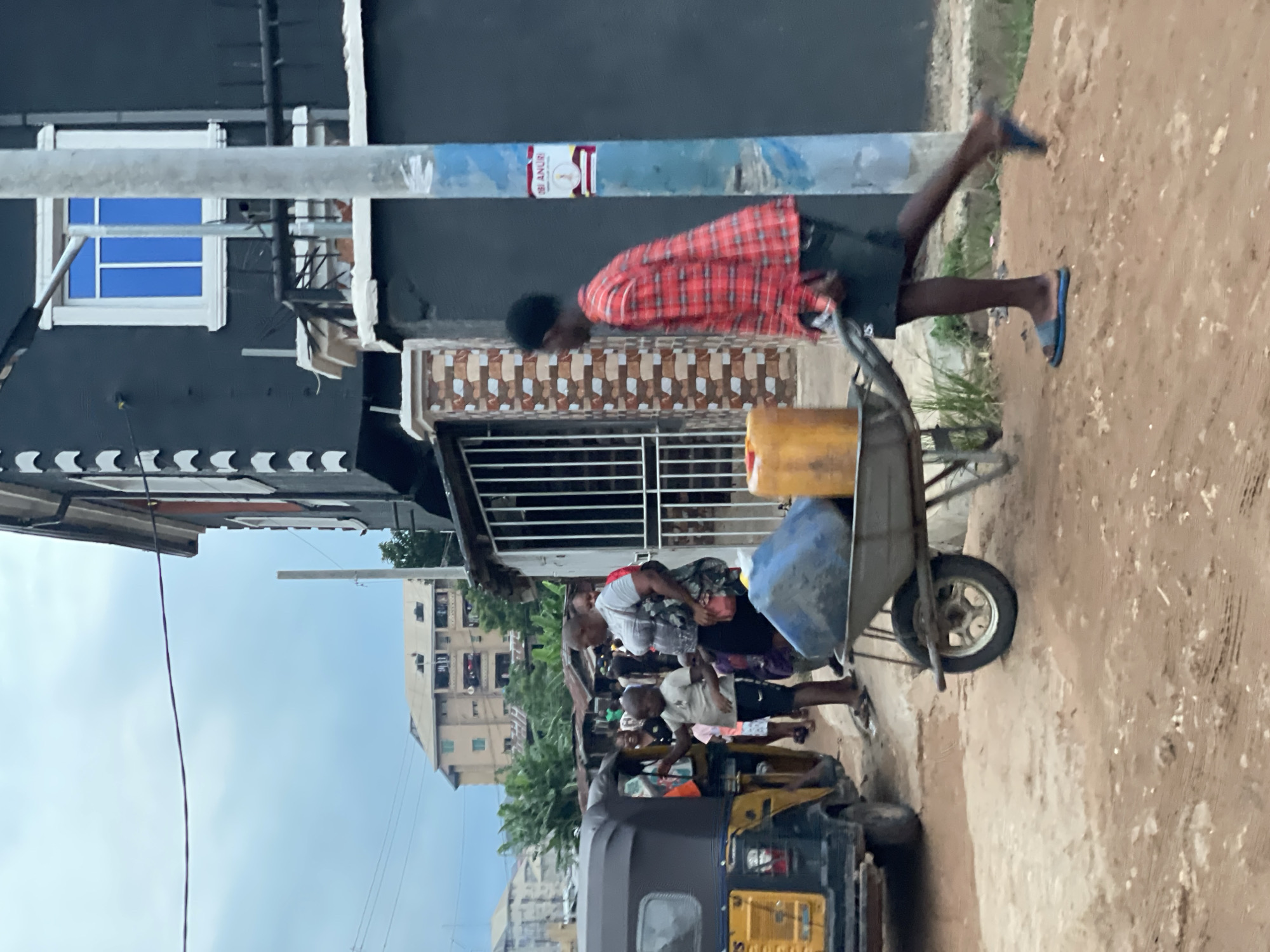 In Nkpor, Nigeria, a man in a red plaid shirt and blue sandals runs along a dirt road, pushing a wheelbarrow containing a yellow jerrycan and a blue container. A crowd of people stands nearby, with an auto-rickshaw parked at the edge of the road. Buildings are visible in the background, including a partially visible dark-colored structure with a white-framed window, and a taller, multi-story apartment building. Overhead power lines are present.