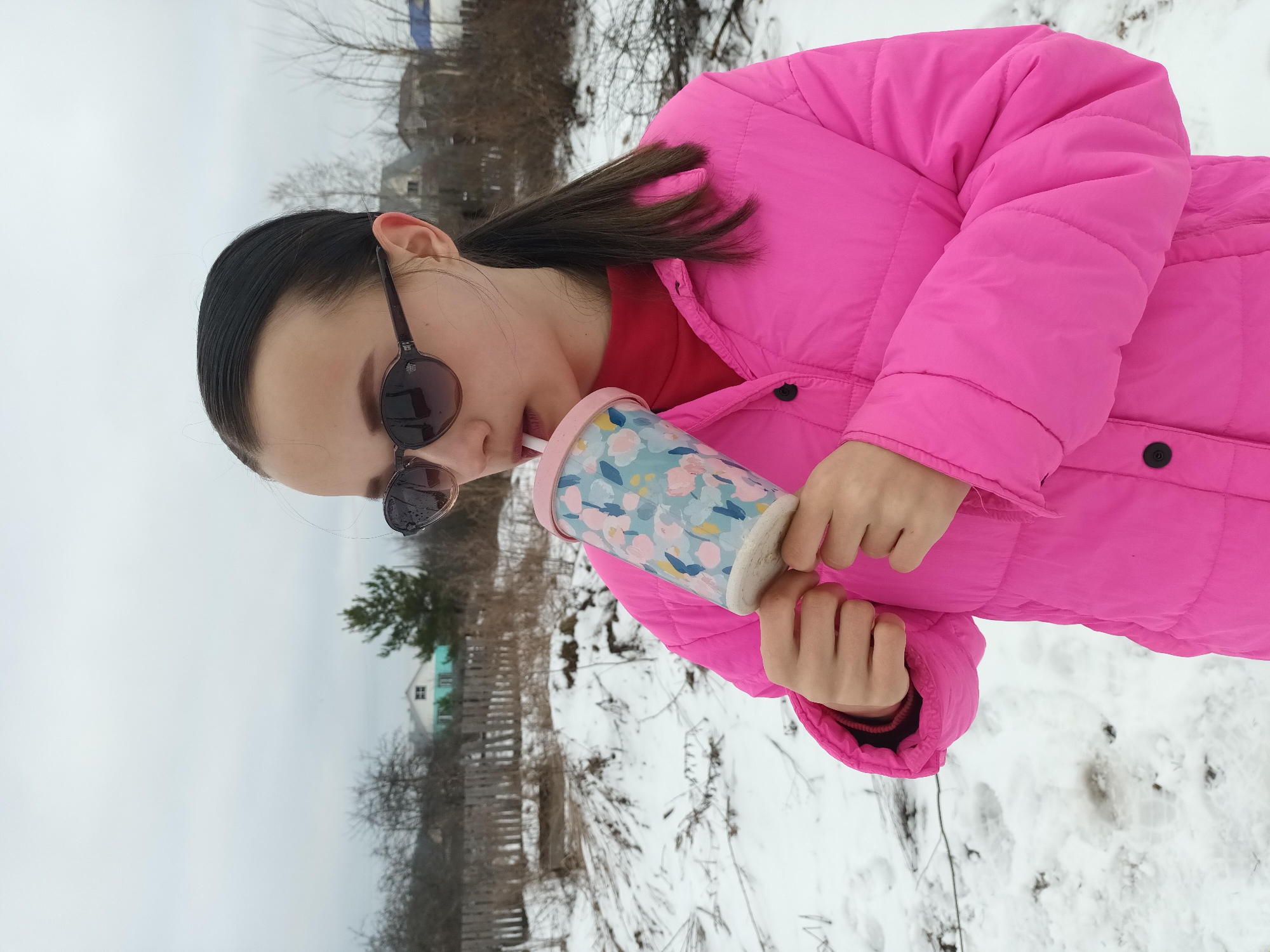 A young woman stands in a snow-covered area, drinking from a floral-patterned tumbler through a straw. She wears a bright pink puffy jacket and sunglasses. In the background, there is a wooden fence, bare trees, and a house with a blue roof visible. The location is identified as Ольховка, Russia.