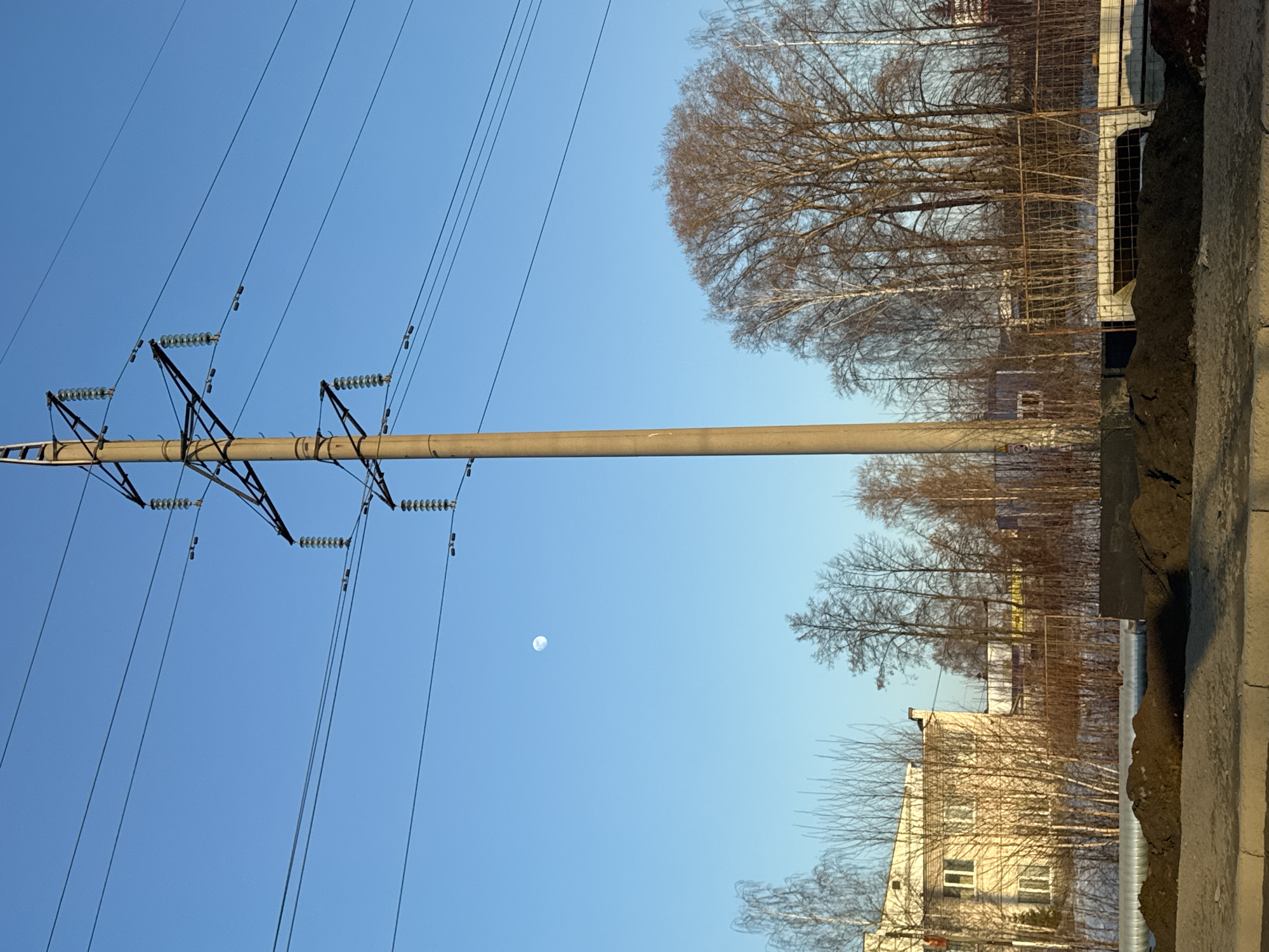 The image shows a sunny day in Chaikovsky, Russia. An electric power transmission pole is in the foreground, with wires running from it against a clear blue sky. A faint, round object, likely the moon, is visible. Beyond the pole, bare deciduous trees line the area. Further in the background, a building with windows is visible. Concrete structures border the lower right of the image, suggesting an urban or suburban setting.