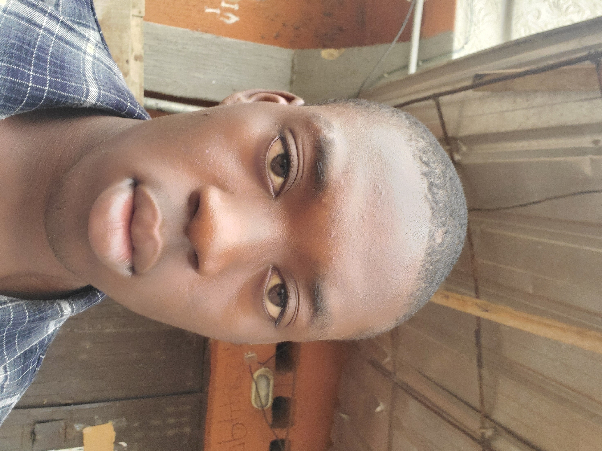 A close-up portrait of a Black male with short, dark hair. He has brown eyes and full lips. He is wearing a blue plaid shirt. The background is blurred and appears to be an outdoor setting with wooden planks and a painted wall visible. The location is Unknown, Unknown.