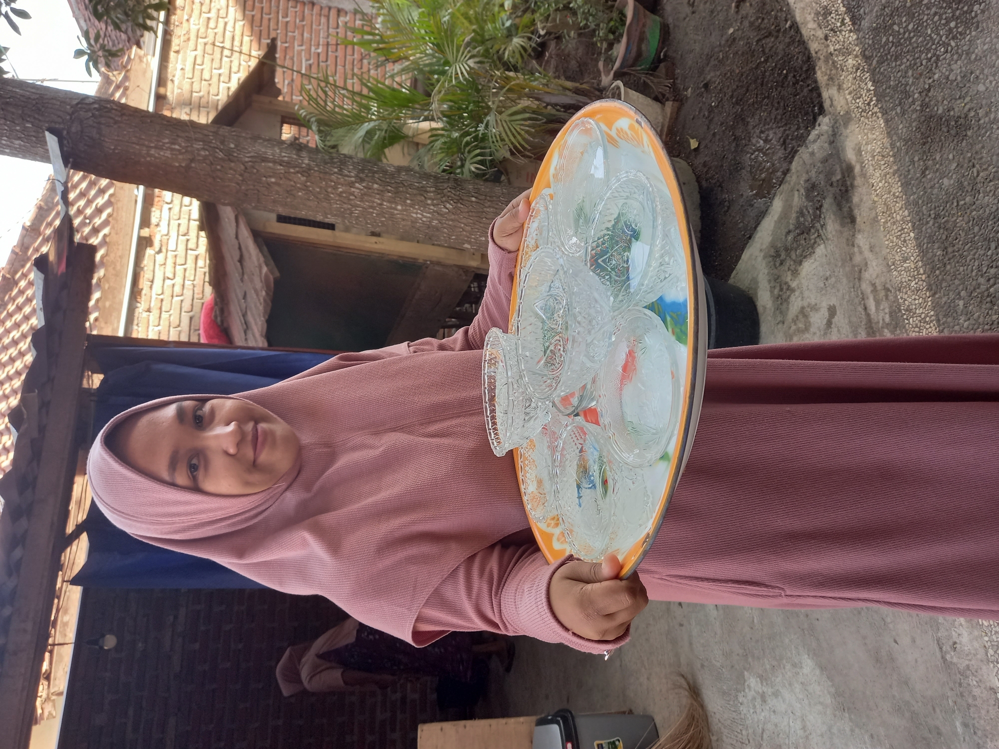 An Indonesian woman in a pink hijab and dress holds a tray of small, clear glass bowls. The bowls appear to be ornate, with textured glass and some containing colorful decorative elements or liquid. She is standing outdoors, with a brick wall and a corrugated metal roof visible in the background. A tree trunk is also prominent in the right side of the frame. The ground is a mixture of concrete and dirt. The setting suggests a residential area.