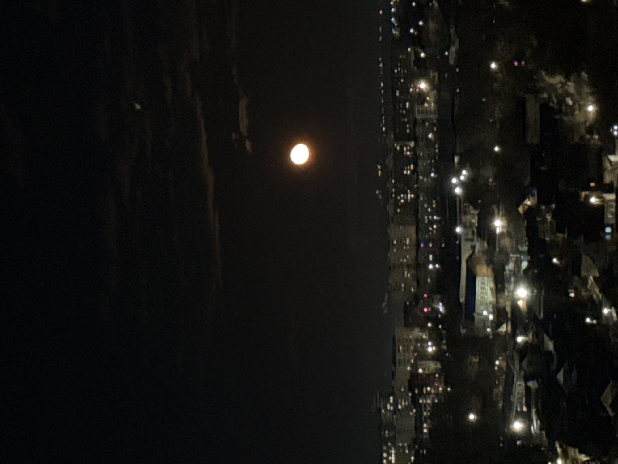 The image depicts a night scene with a full moon dominating the dark sky. To the right of the moon is a cityscape with numerous illuminated windows and streetlights indicating urban activity. The location appears to be Izhevsk, Russia, based on the building style and layout. The foreground is predominantly dark, with subtle gradations suggesting clouds or atmospheric haze. No people are directly visible.