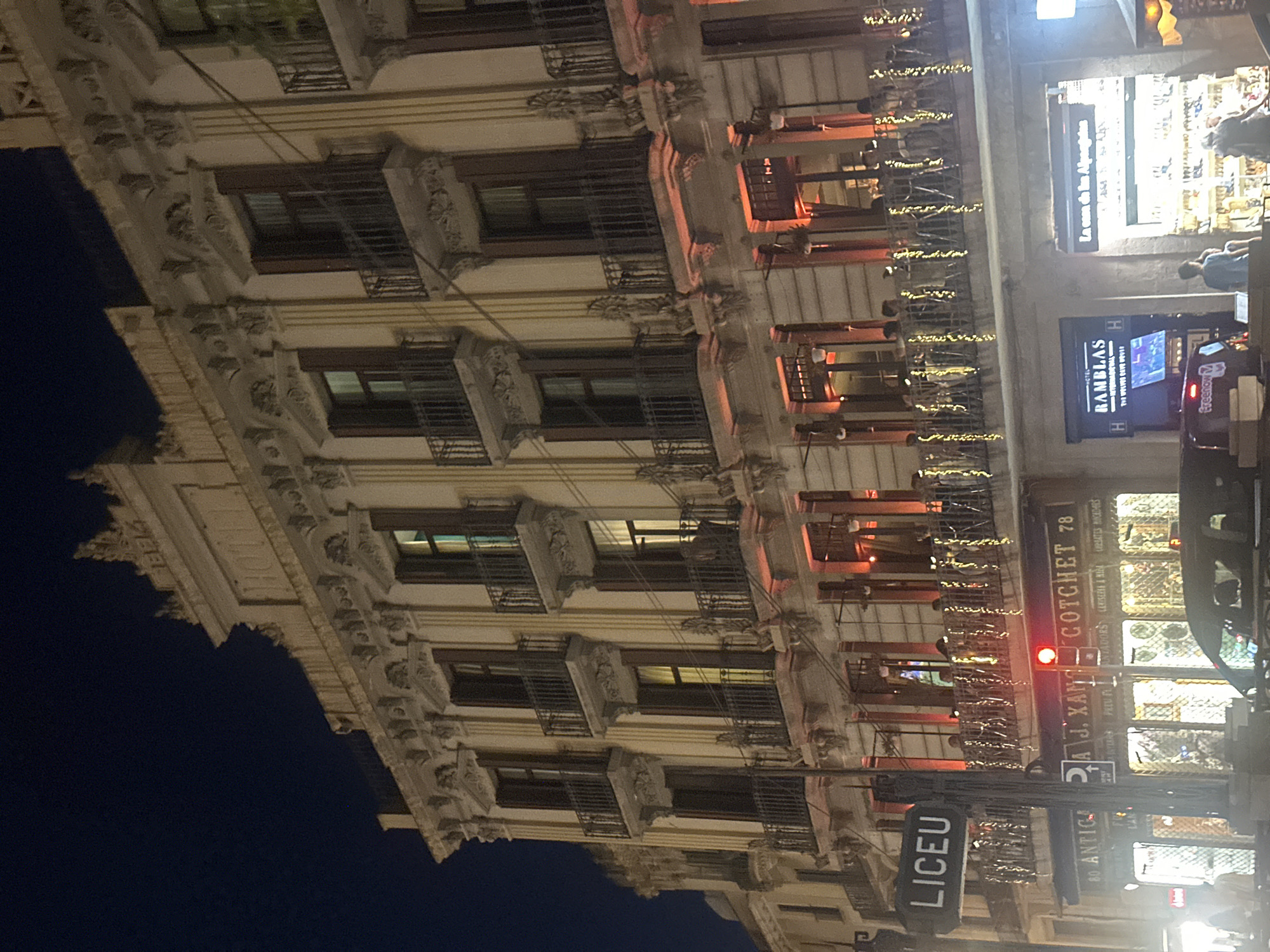 The image shows a multi-story building on a street at night in Barcelona, Spain. The building is ornate, with detailed architectural features, including balconies with dark railings and numerous windows. Some windows are lit from within, casting a warm glow. Red lights illuminate the lower facade of the building. String lights are draped along the railing of a balcony on the second level.
On the ground level, several storefronts are visible. A sign with the word "LICEU" is mounted on a pole on the left. To the right of the "LICEU" sign, there are shops with illuminated displays and signage, including "ANTIGA J. XAN GOTCHET 78," "HOTEL RAMBLAS," and "La casa de los Alpentinos." People are visible walking on the sidewalk and inside some of the establishments. A traffic light with a red signal is also present. The overall impression is of a lively urban scene at night.