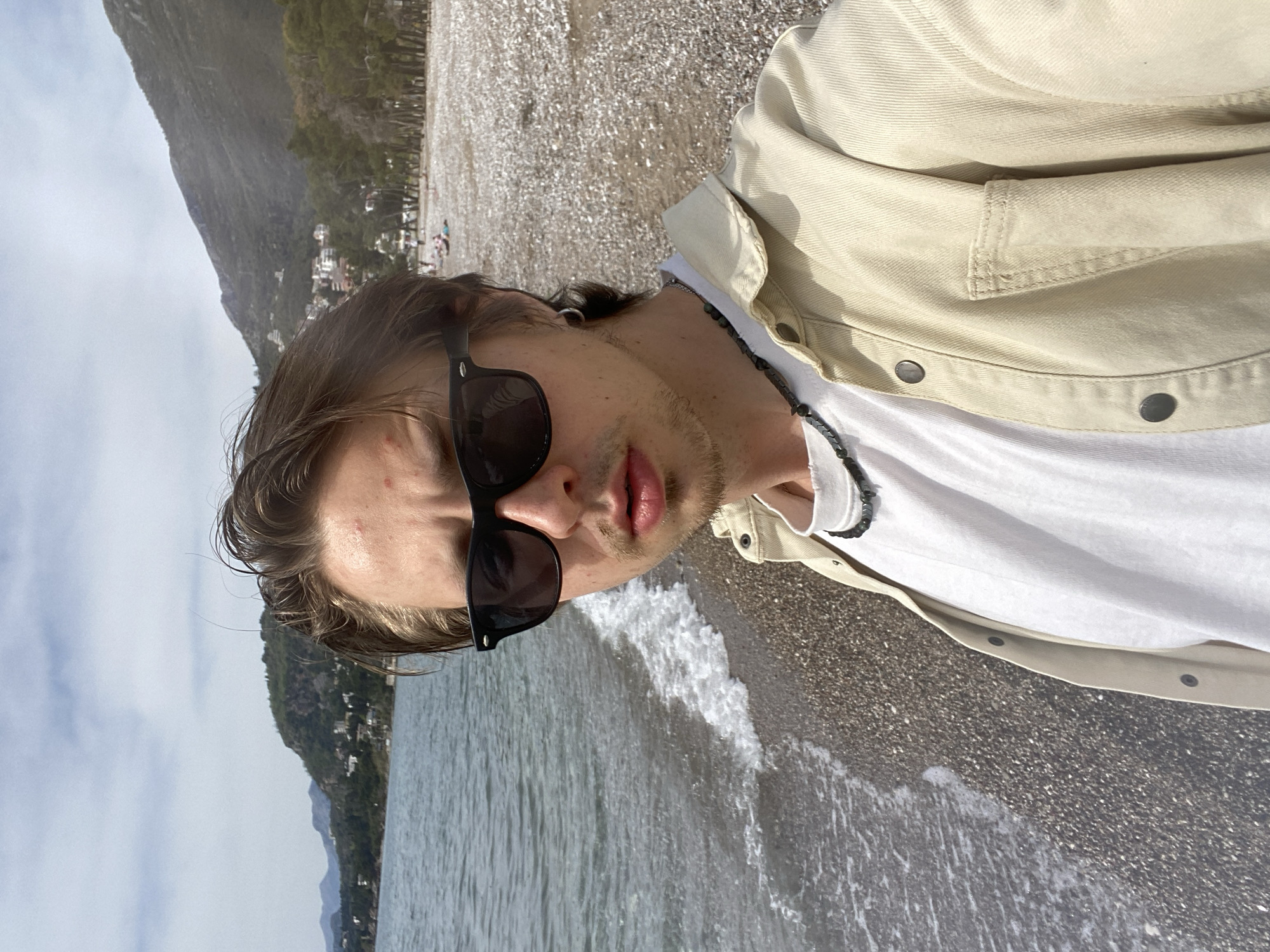 A selfie of a light-skinned male wearing a white t-shirt, light beige jacket, and dark sunglasses, taken on a pebbly beach in Bar, Montenegro. The man has short, wet hair and a light stubble. A beaded necklace is visible around his neck. The ocean water is lapping at the shore. A mountain covered in trees and buildings is in the background, with a cloudy sky. The man is standing at the edge of the water.