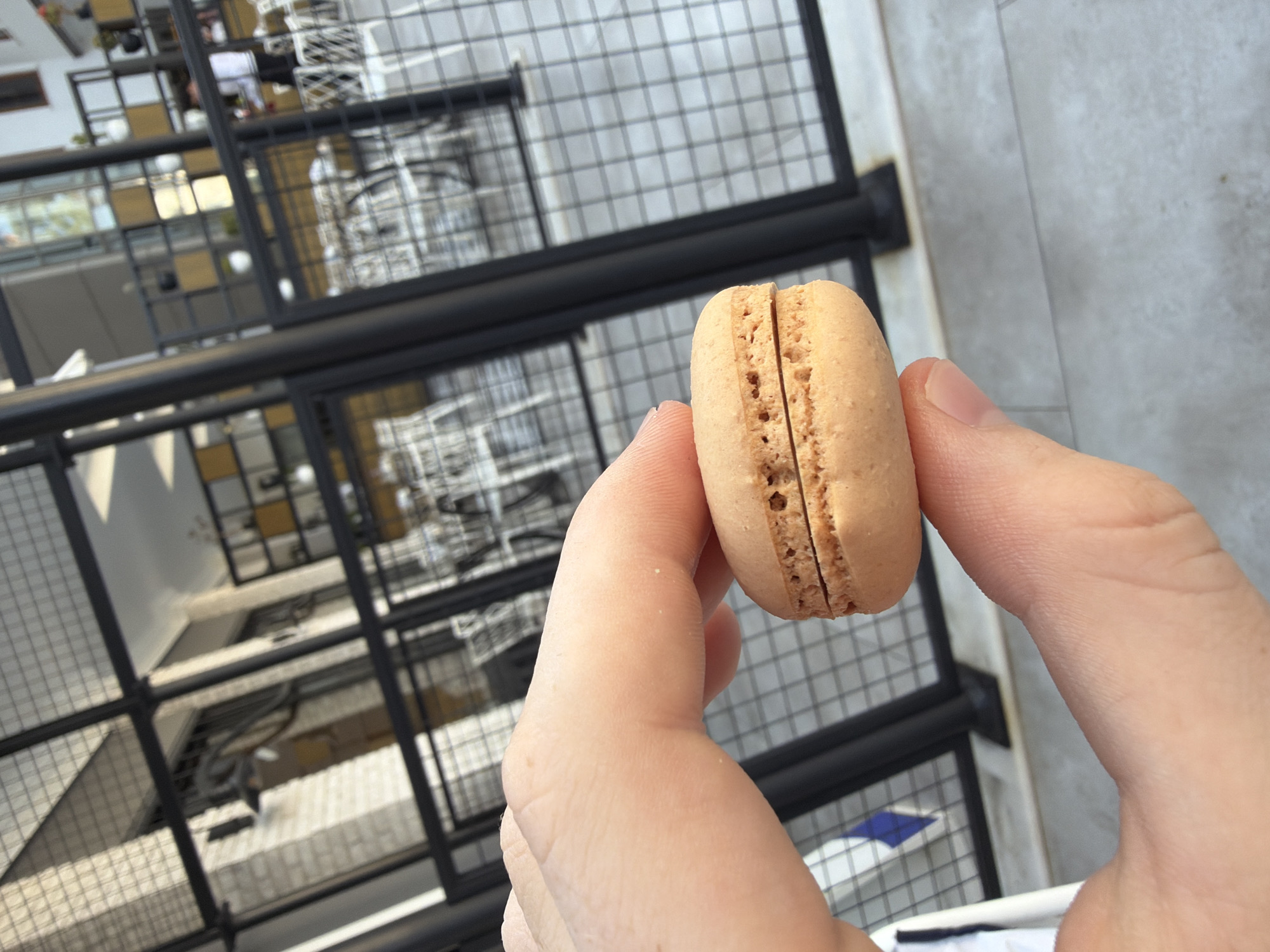 In Kemer, Turkey, a close-up shot captures a hand holding a single, light brown macaron with a distinct visible filling. The macaron rests between the thumb and forefinger, with the textured surface of the cookie clearly visible, including its characteristic ruffled edge and airy interior. The background is a blur of an outdoor setting, featuring a black metal fence with a diamond grid pattern. Beyond the fence, what appears to be a cafe or restaurant setting is discernible, with tables and chairs arranged. The overall lighting suggests it is daytime, possibly outdoors in an urban environment.