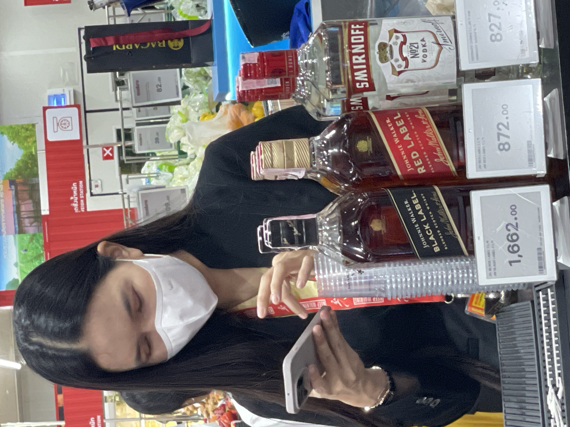 In Pattaya City, Thailand, a woman wearing a white face mask and a black jacket is holding a smartphone and looking at it. She is standing in front of a display of alcoholic beverages, including bottles of Johnnie Walker Red Label and Smirnoff Vodka. In the background, shelves are stocked with various items, and there is a red sign with Thai text and a weighing scale icon. A black Bacardi bag is also visible. Price tags are displayed for the alcoholic beverages, showing prices such as 1,662.00, 872.00, and 827.00. Plastic cups are stacked next to the bottles. The lighting suggests an indoor retail environment, possibly a supermarket or duty-free shop.